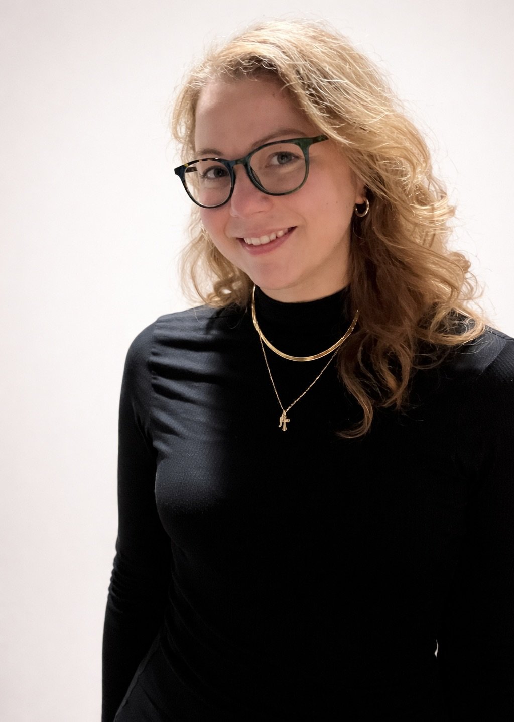 Portrait of a woman with blonde hair wearing glasses, earrings, layered necklaces, and a black turtleneck, smiling against a plain white background.