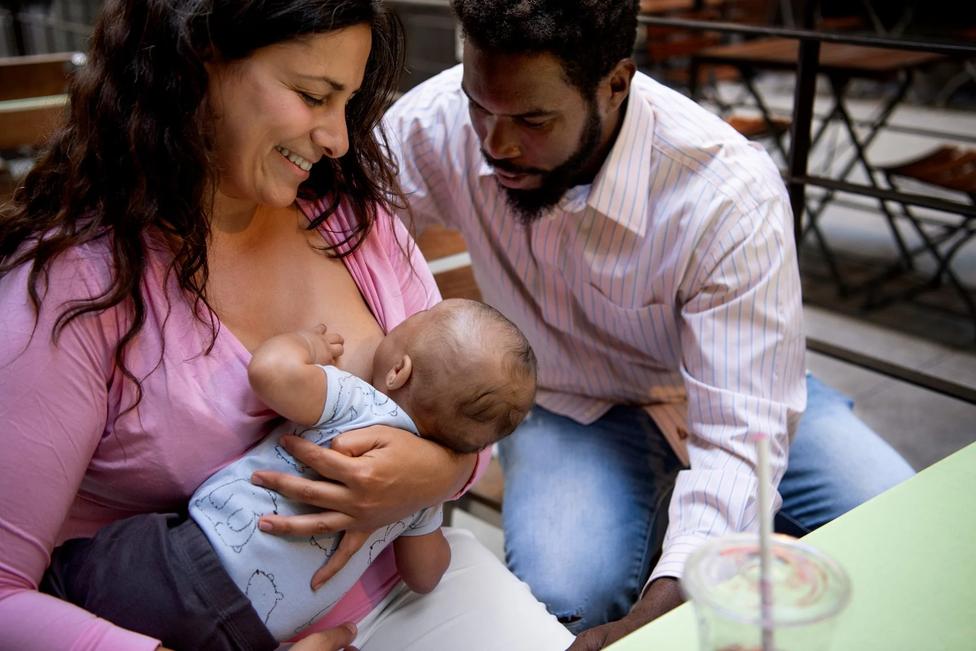 A woman nursing a baby while a man looks at the baby; they are sitting outdoors at a table with a drink in the foreground.