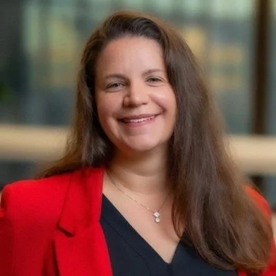 Portrait of a woman with long brown hair, wearing a red blazer and a black top, smiling indoors with blurred windows in the background.