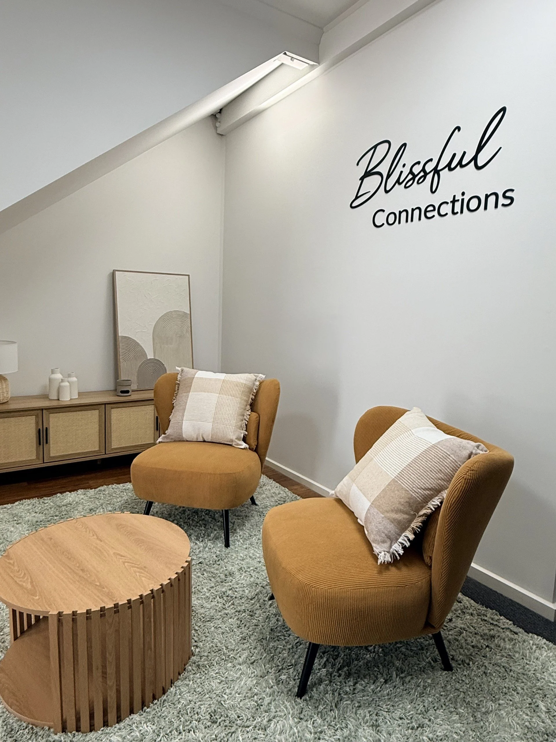 Blissful Connections consulting room, Cozy waiting or lounge area with two mustard yellow armchairs with beige and plaid cushions, a wooden coffee table, a carpet, and a sideboard with decorative items. Wall sign reads 'Blissful Connections'.