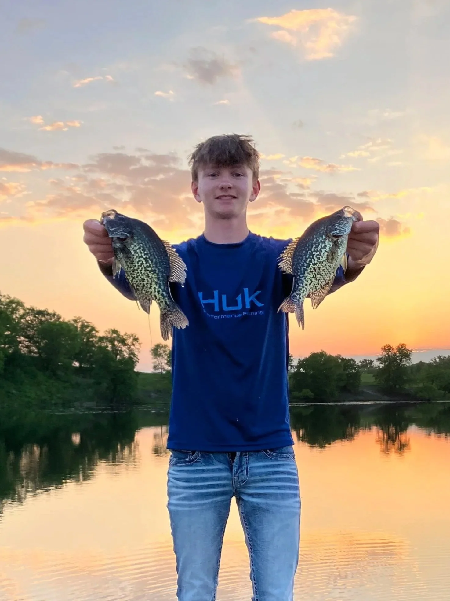A young man holding two fish, one in each hand, outdoors near a body of water during sunset.