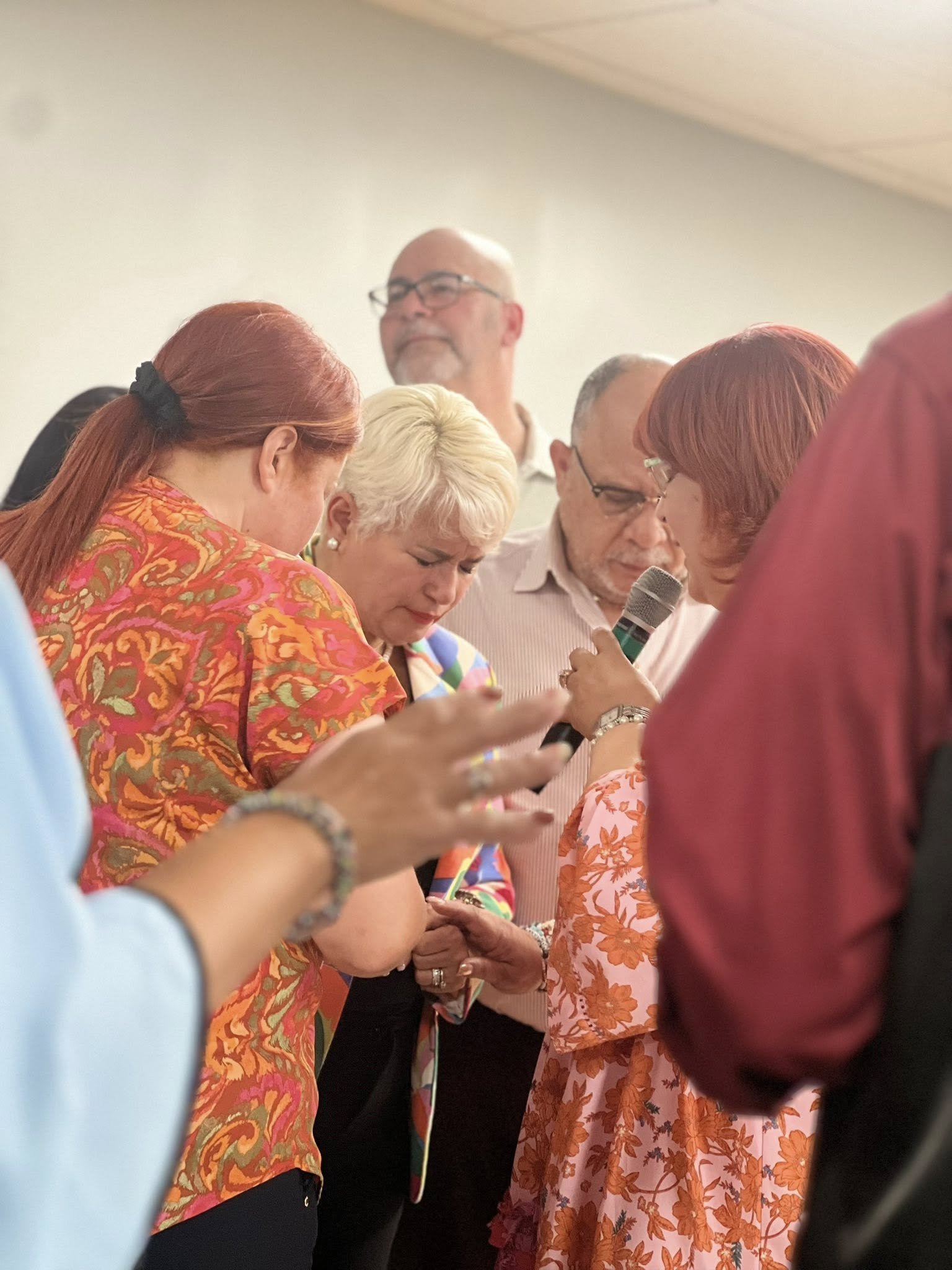Group of people praying together at an indoor gathering, with some holding a microphone and looking down.