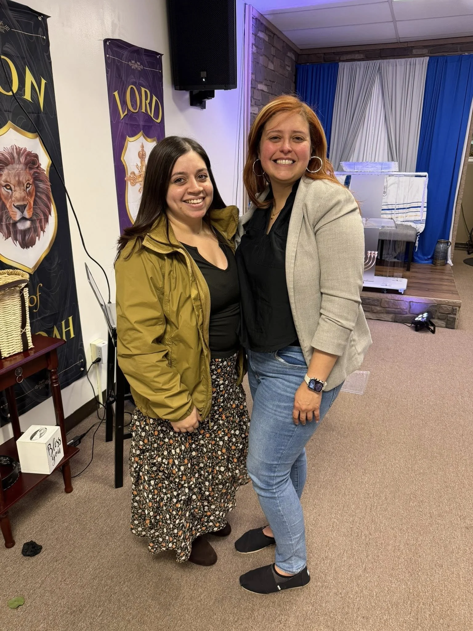 Two women standing close together inside a church, smiling at the camera. One woman wears a green jacket and floral skirt, the other wears a beige blazer and jeans. Behind them are church banners and a pulpit with a purple and white curtain.
