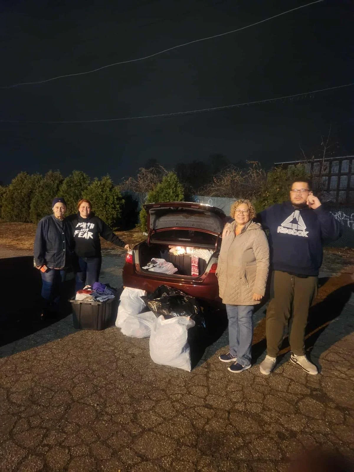 Four people standing outdoors at night next to an open car trunk with bags of items, some on the ground, in a rural area with trees and power lines visible.