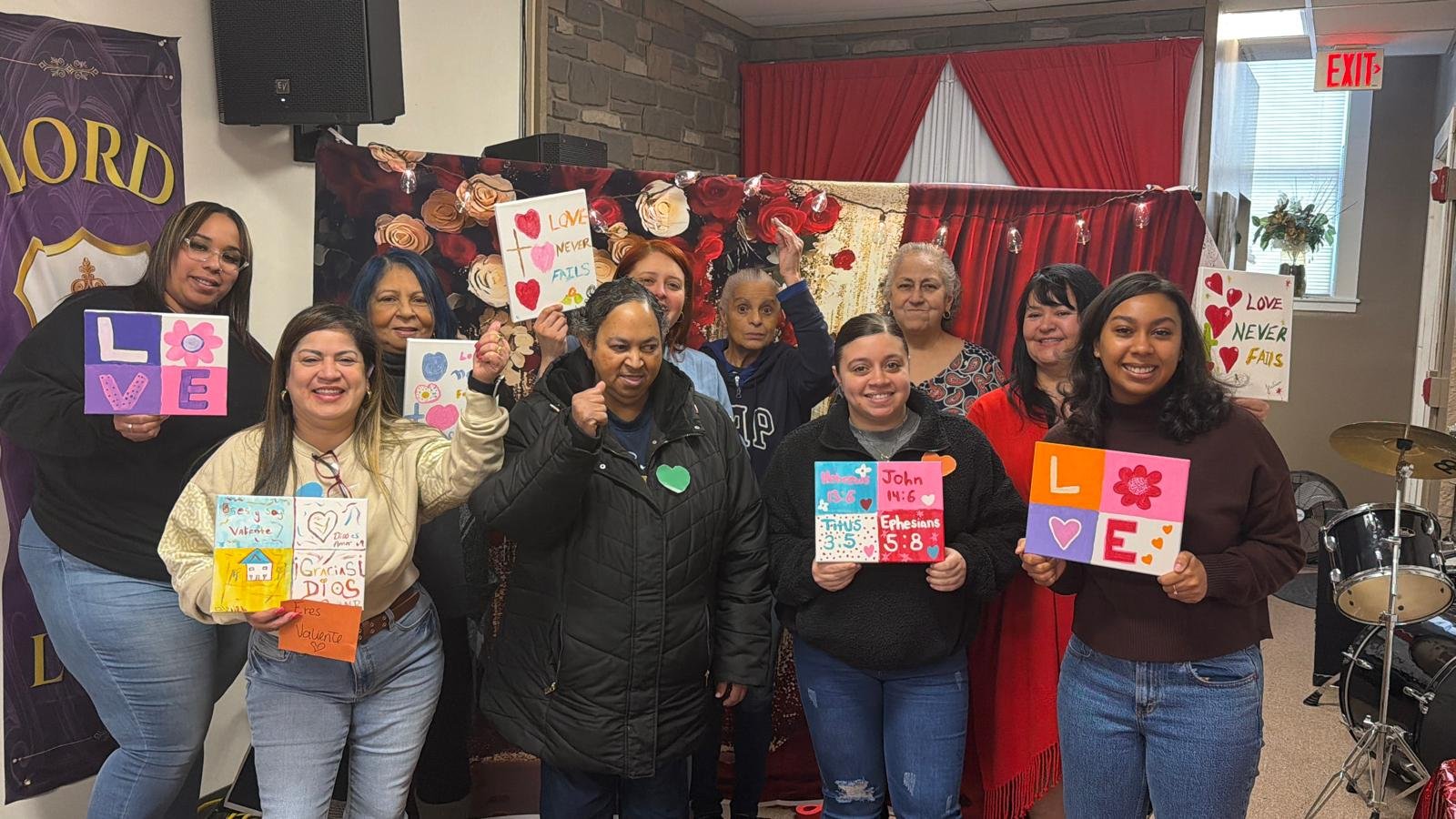 A group of women smiling and holding colorful handmade signs and posters with messages about love and faith in a community center, with a decorated red curtain backdrop and drums on the side.
