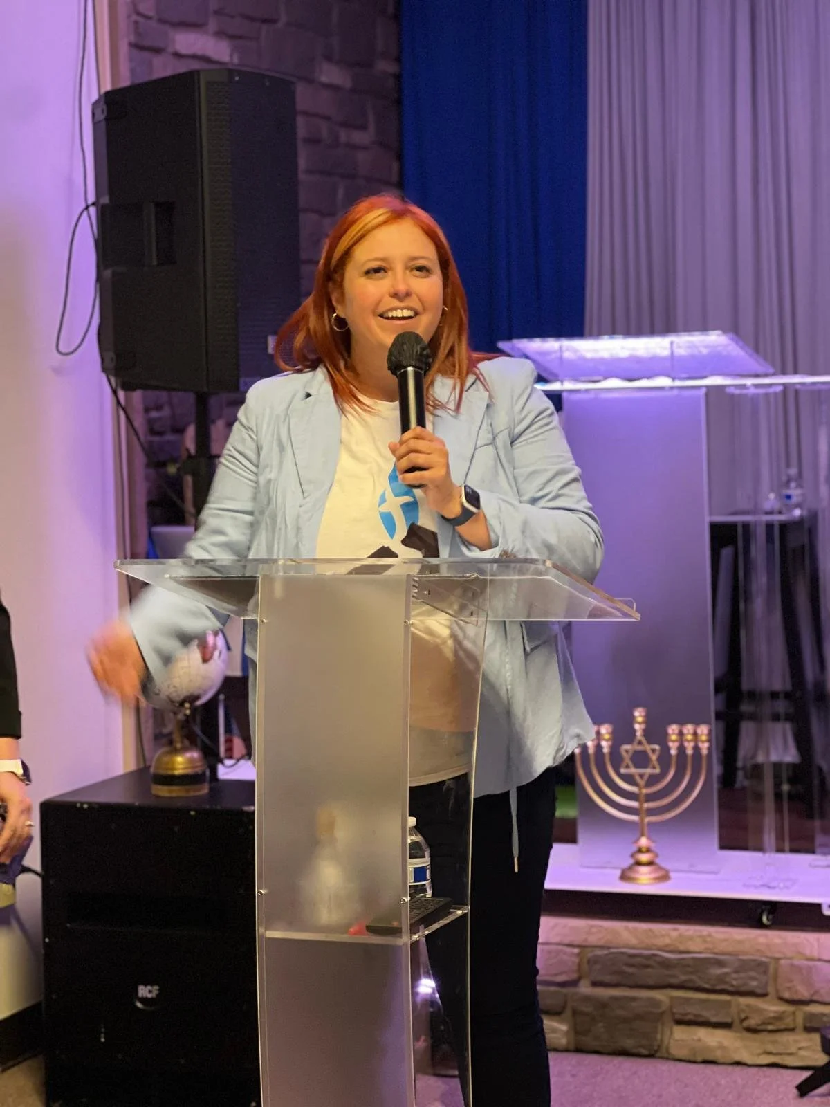 A red-haired woman speaking into a microphone at a lectern with a menorah and a globe in the background.