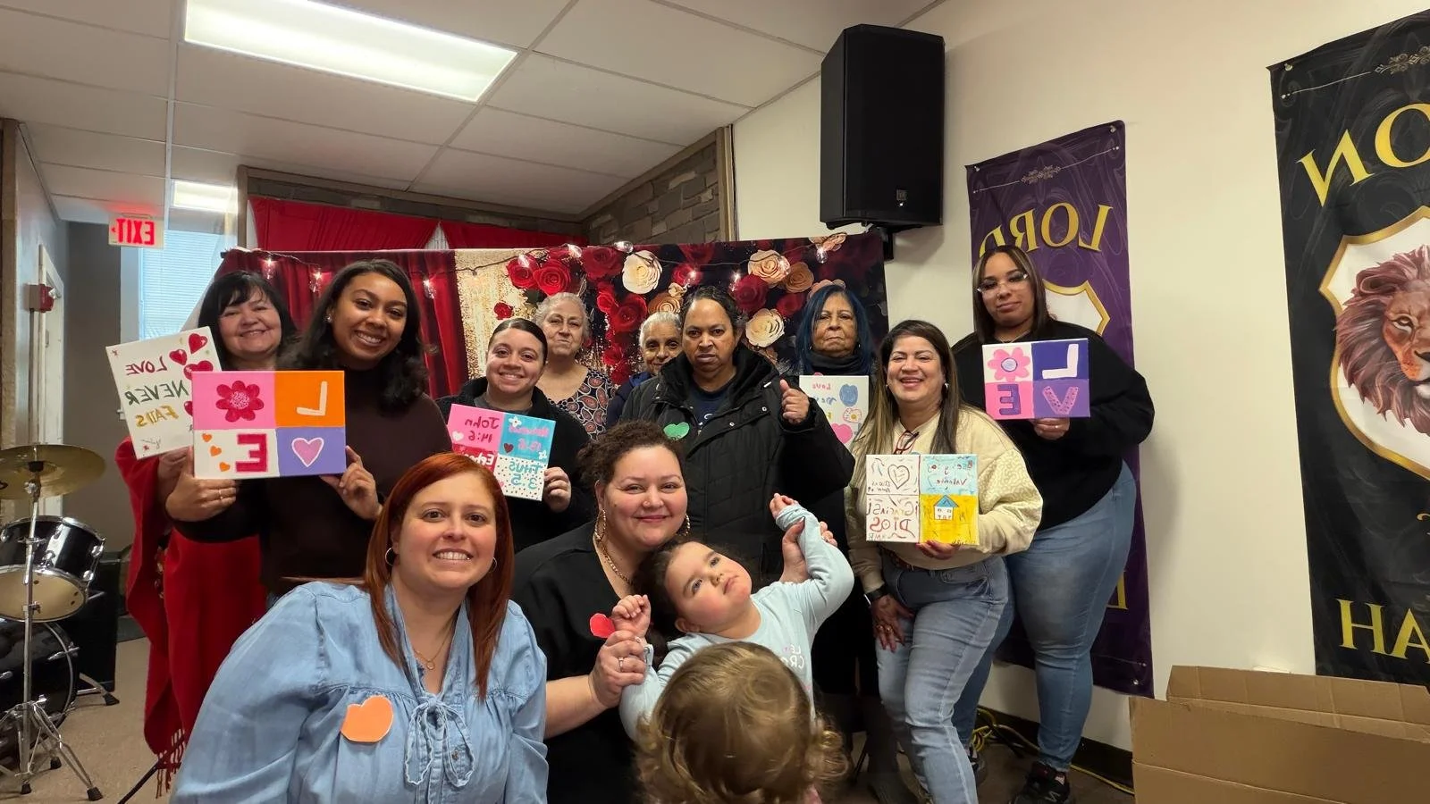 Group of women and children celebrating Valentine's Day, holding handmade cards with love messages, in a decorated room with banners and a red curtain in the background.