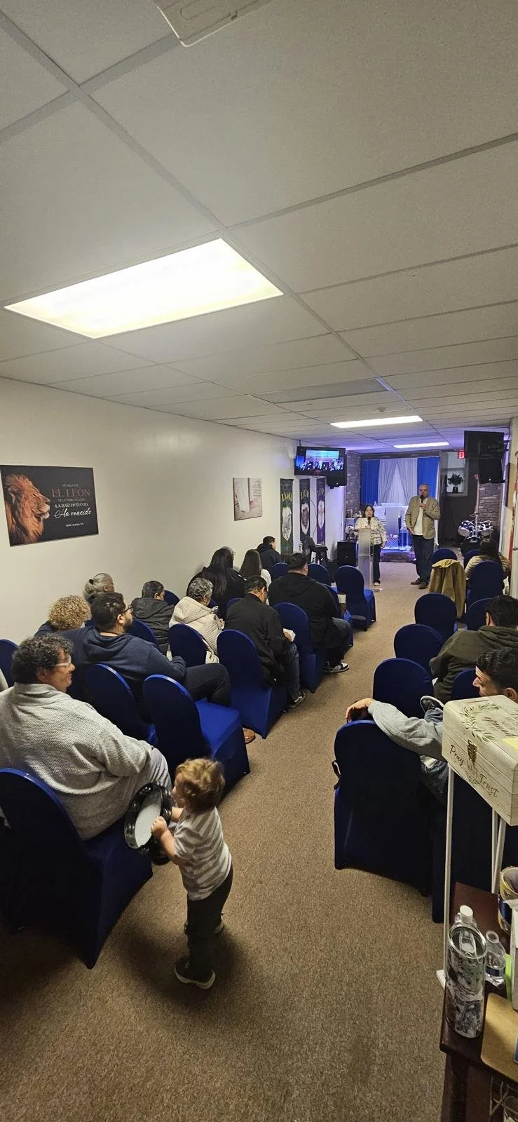 A group of people seated on blue chairs in a small room listening to a speaker at a podium. There is a small child holding a tambourine near the front.