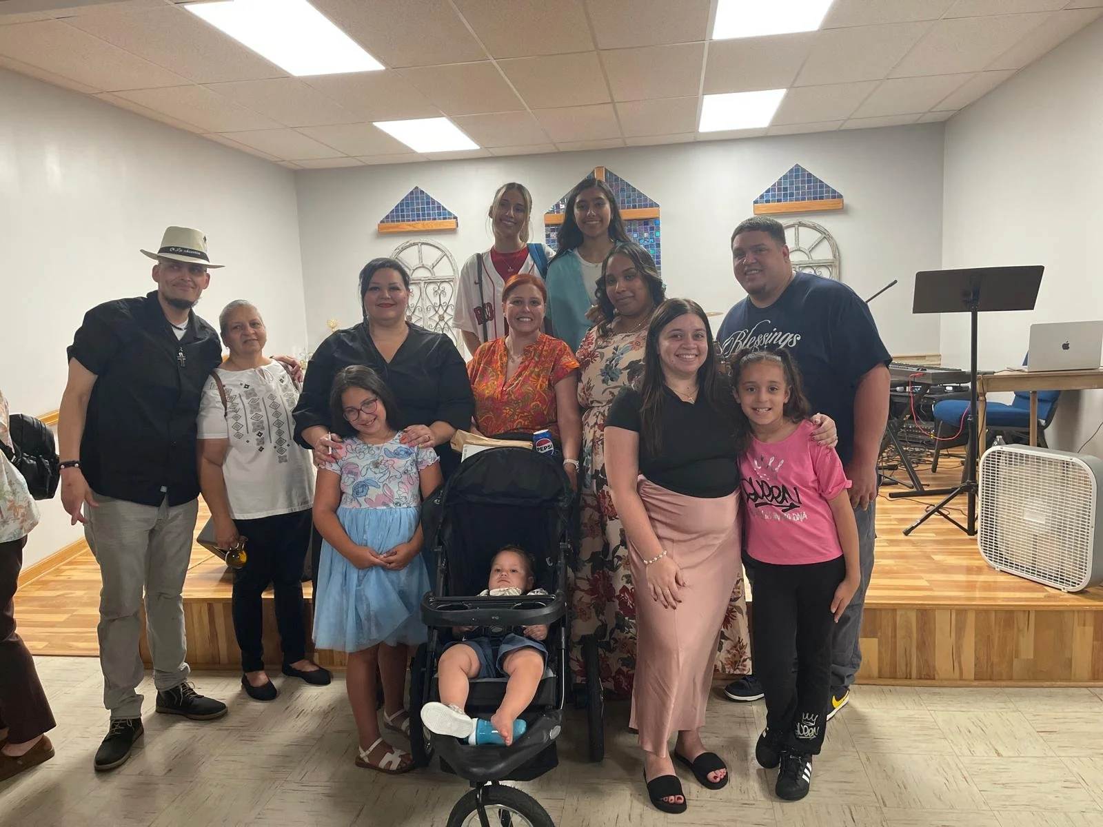 A group of people, including children and adults, poses for a photo in a room with a wooden stage and stained glass windows in the background. Some are smiling and standing, while children sit or are held in front.