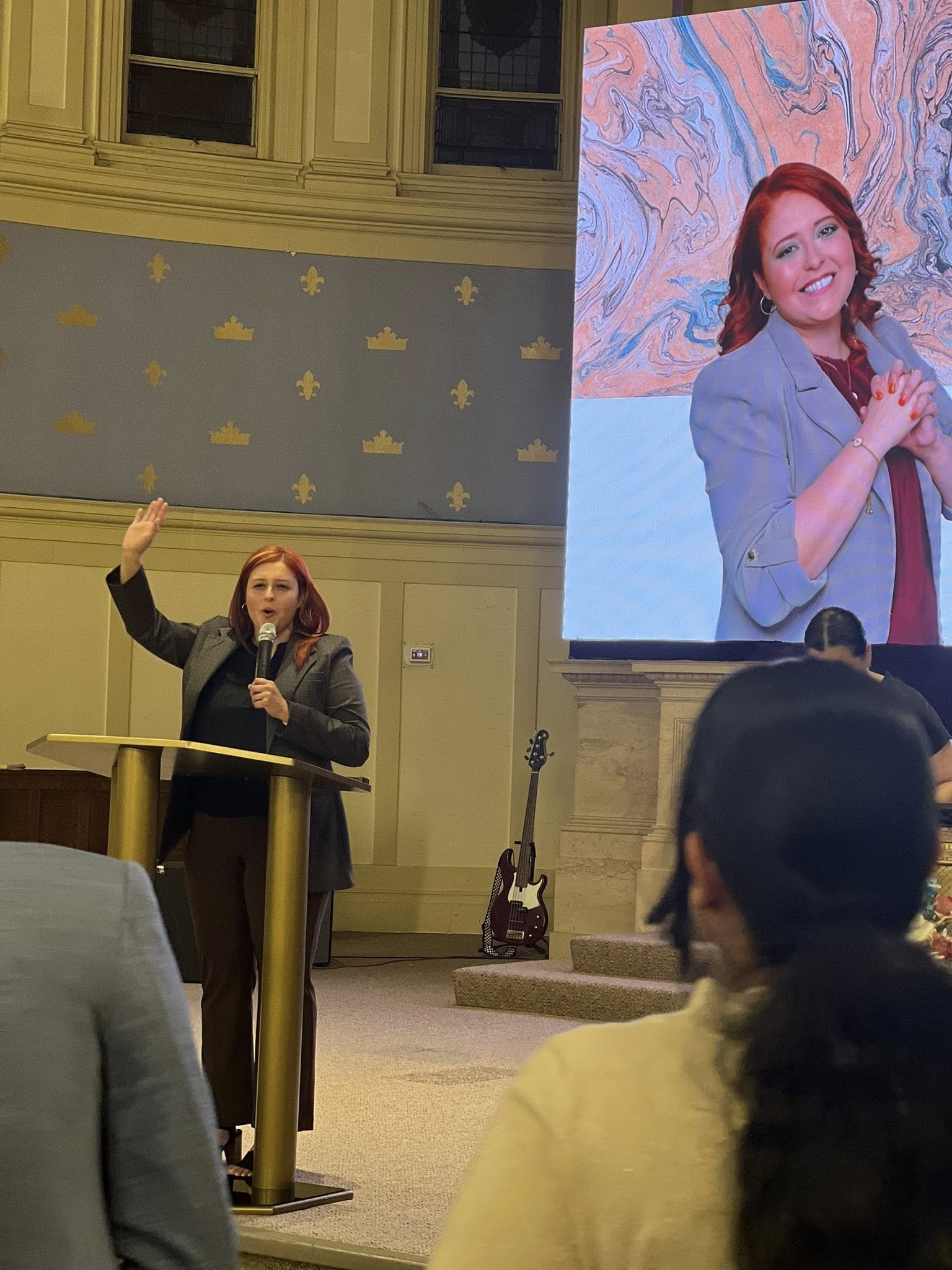 A woman with red hair speaking at a podium during a presentation or speech, with a large screen behind her showing her portrait. There are people seated in the audience, and musical instruments are visible to the side of the stage.