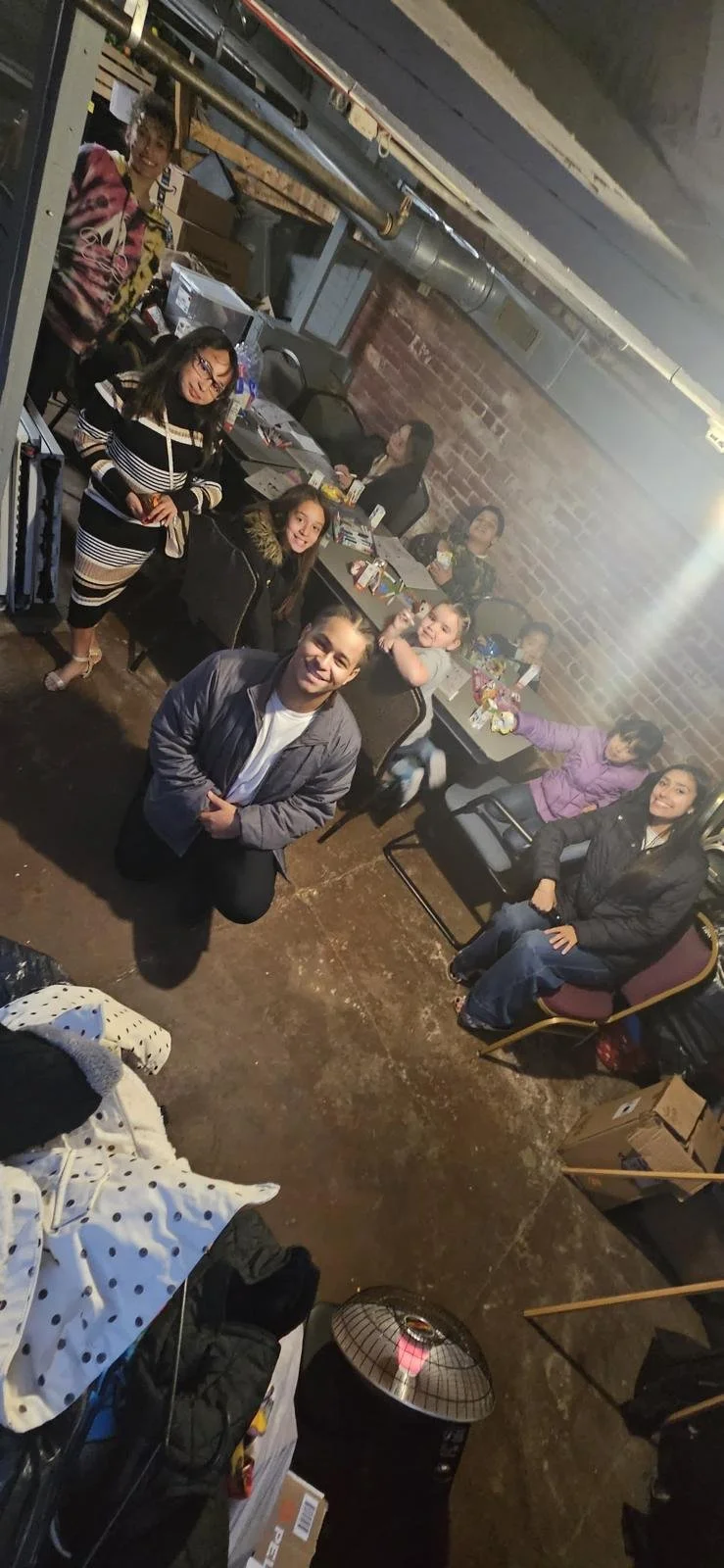 A group of people sitting around tables in a basement or industrial-style room with exposed brick walls and pipes, some smiling and looking at the camera, with snacks and drinks on the tables.