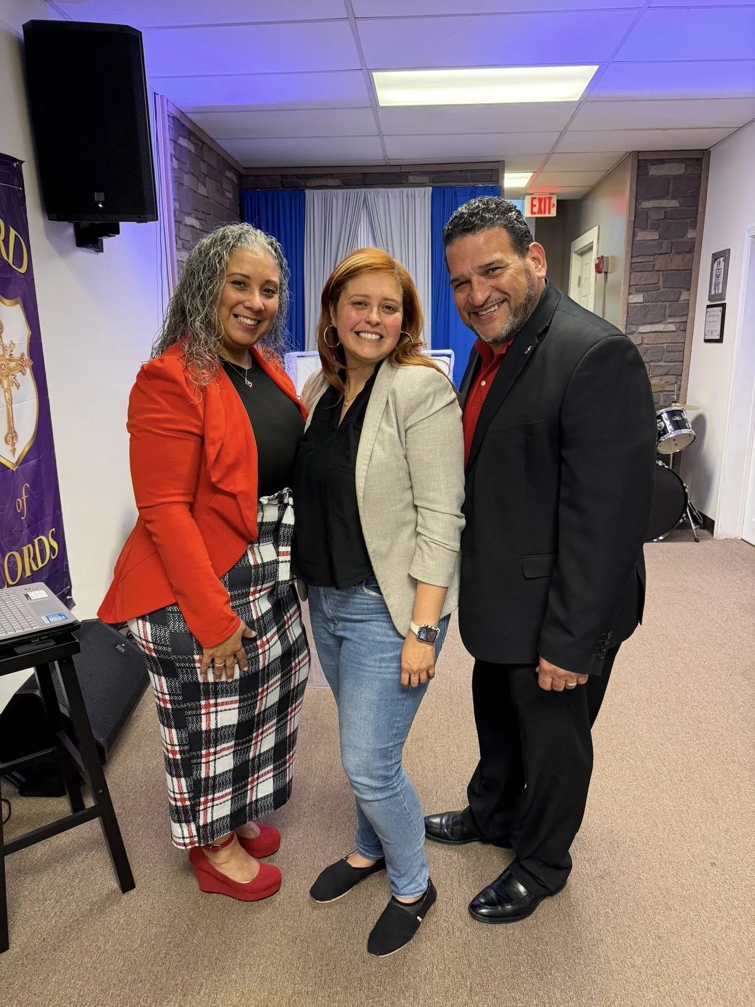 Three people standing together, smiling, at an event or meeting. The woman on the left has gray curly hair, wearing a red blazer, black top, plaid skirt, and red shoes. The woman in the middle has red hair, wearing a beige blazer, black top, jeans, a