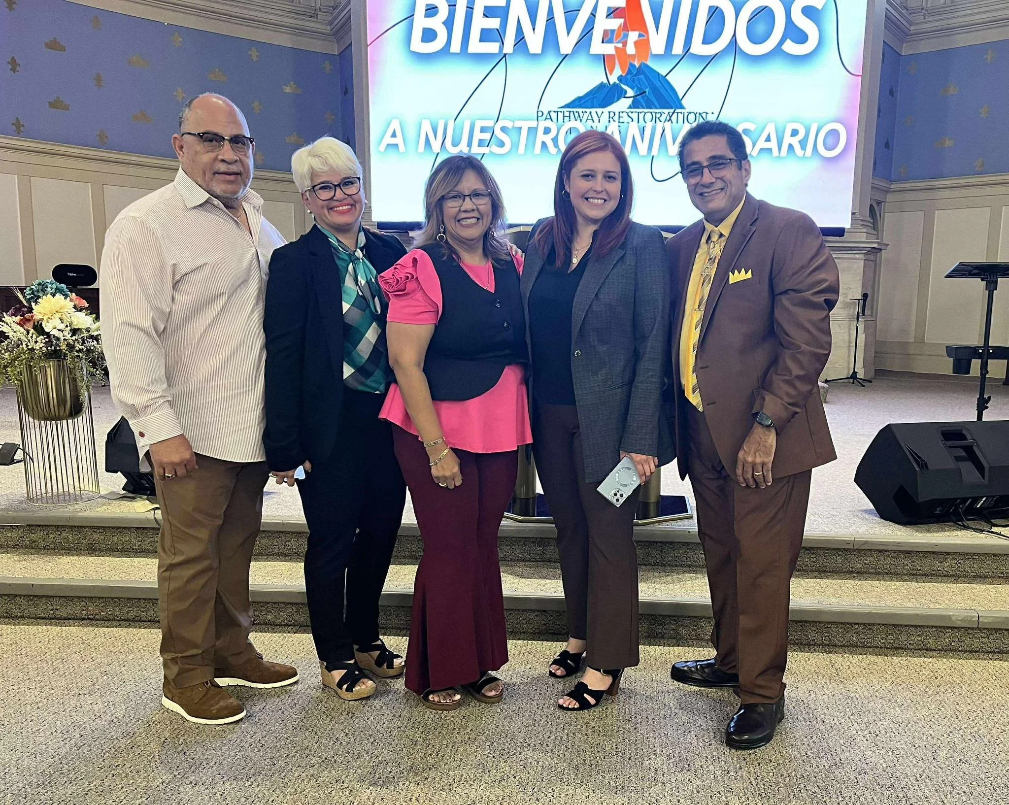 Five people standing on a stage, smiling for the camera. Behind them is a large screen with Spanish words 'BIENVENIDOS' and other text, along with a graphic of a mountain and a sun.