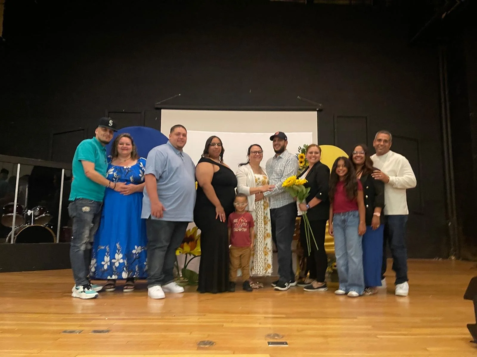 Group of people on stage, some holding flowers, celebrating with a woman in a white floral dress, children, and a man in a white shirt, against a backdrop with sunflowers and decorative elements.