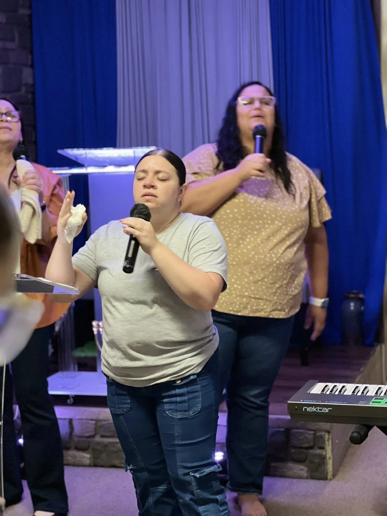 Three women singing in a karaoke setting, with one woman in the foreground holding a microphone and eyes closed, with a keyboard visible on the right.