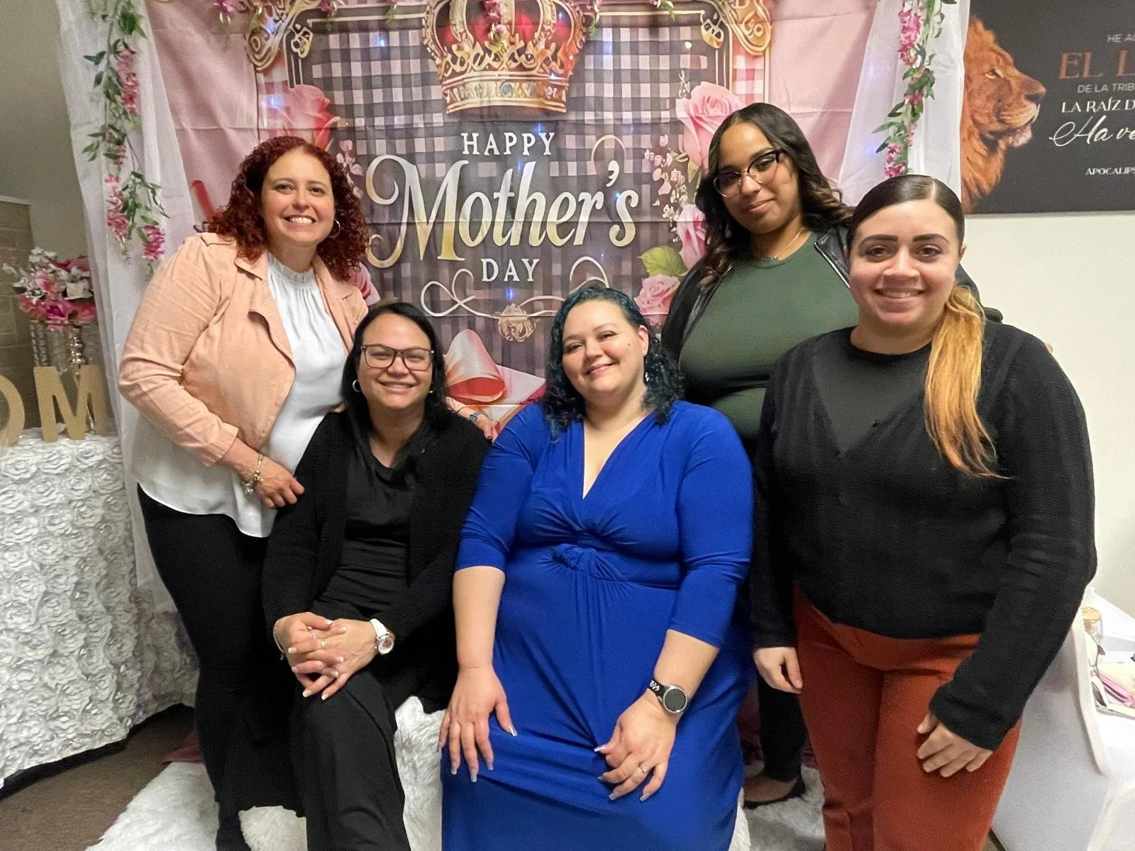 Six women gathered together for a photo in front of a Mother's Day themed decorations backdrop, smiling and posing for the camera.