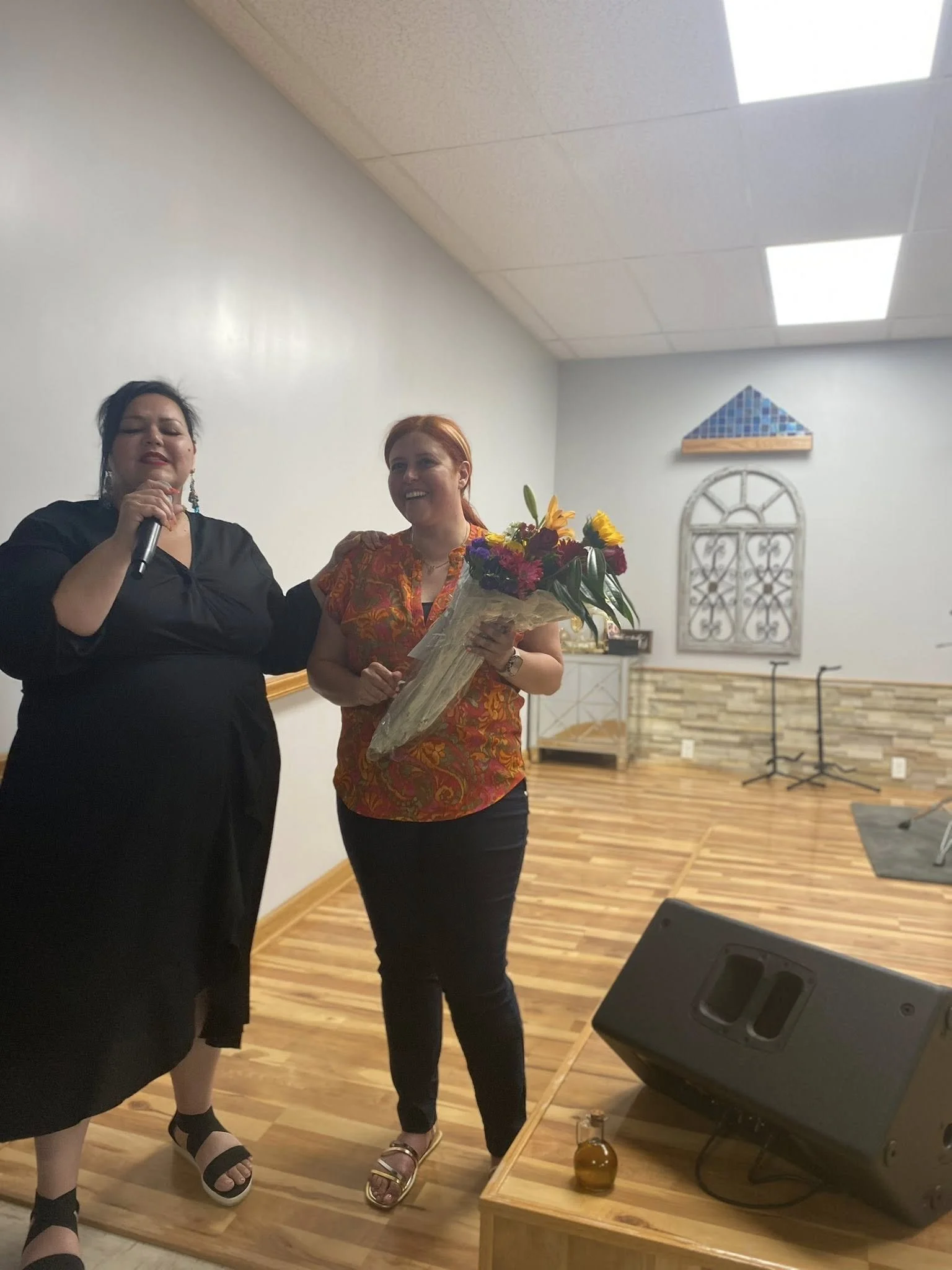 Two women standing together on a stage, one holding a microphone and the other holding a bouquet of flowers, smiling. The stage has a wooden floor and a white wall with decorative elements in the background.