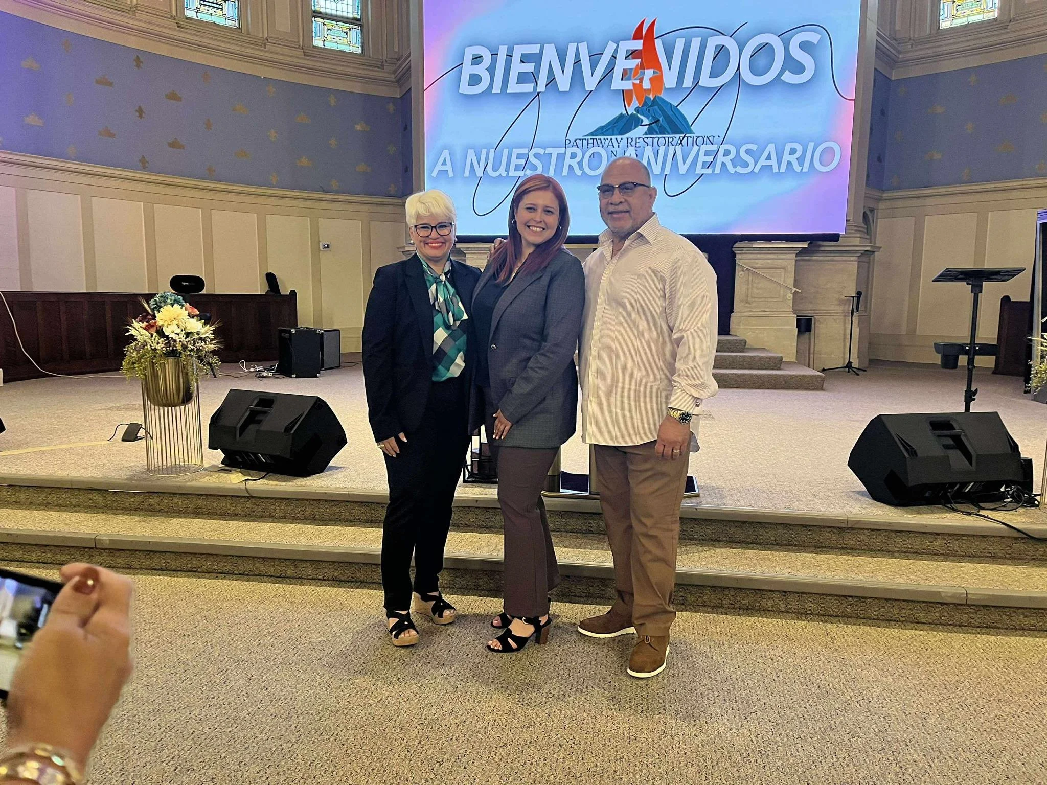 Three people standing on a stage in front of a large screen that says in Spanish "Bienvenidos A Nuestro Aniversario" (Welcome to Our Anniversary). The stage is decorated with flowers and there are speakers and microphones visible.