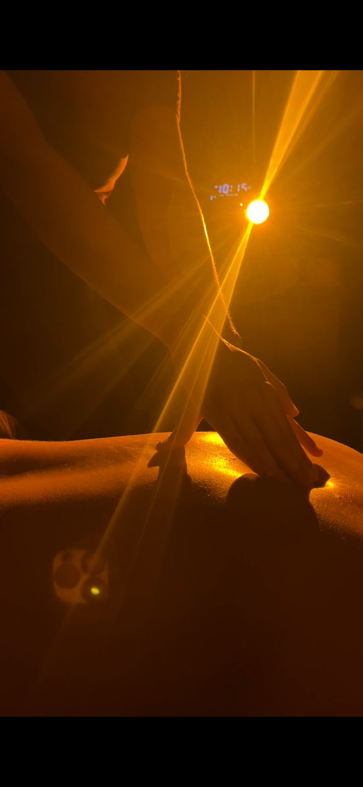 A person receiving a massage in warm, golden lighting, with a visible clock showing 1:10.