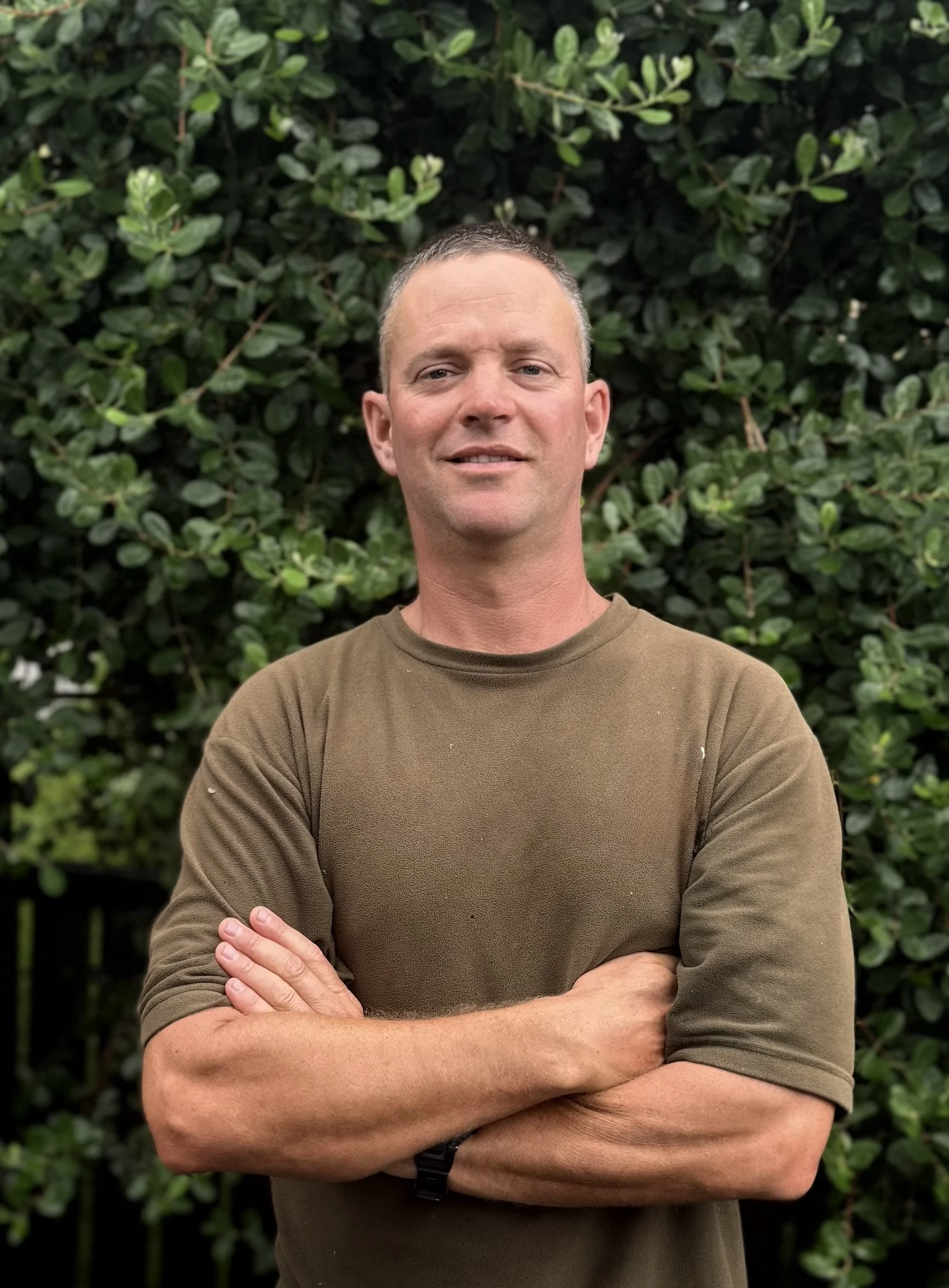 A builder specialised in gutter conversion with short hair and a slight smile standing outdoors with arms crossed in front of dense green foliage.