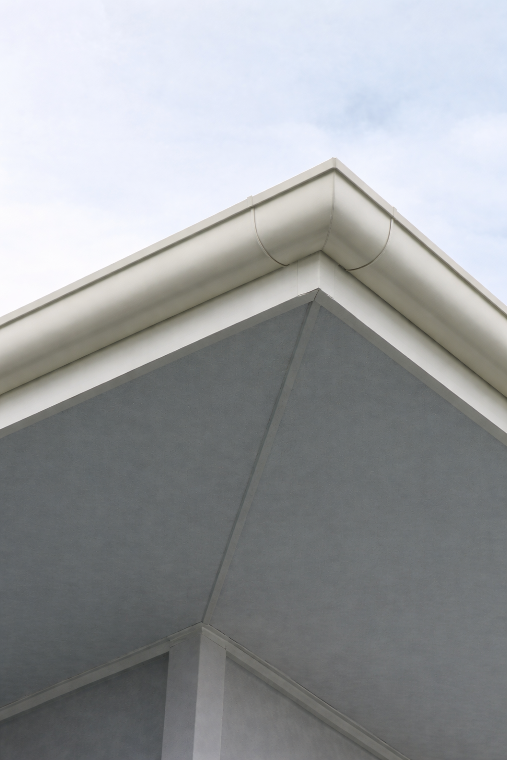 Close-up of the corner of a modern building with a white external gutter and gray soffit, against a cloudy sky.