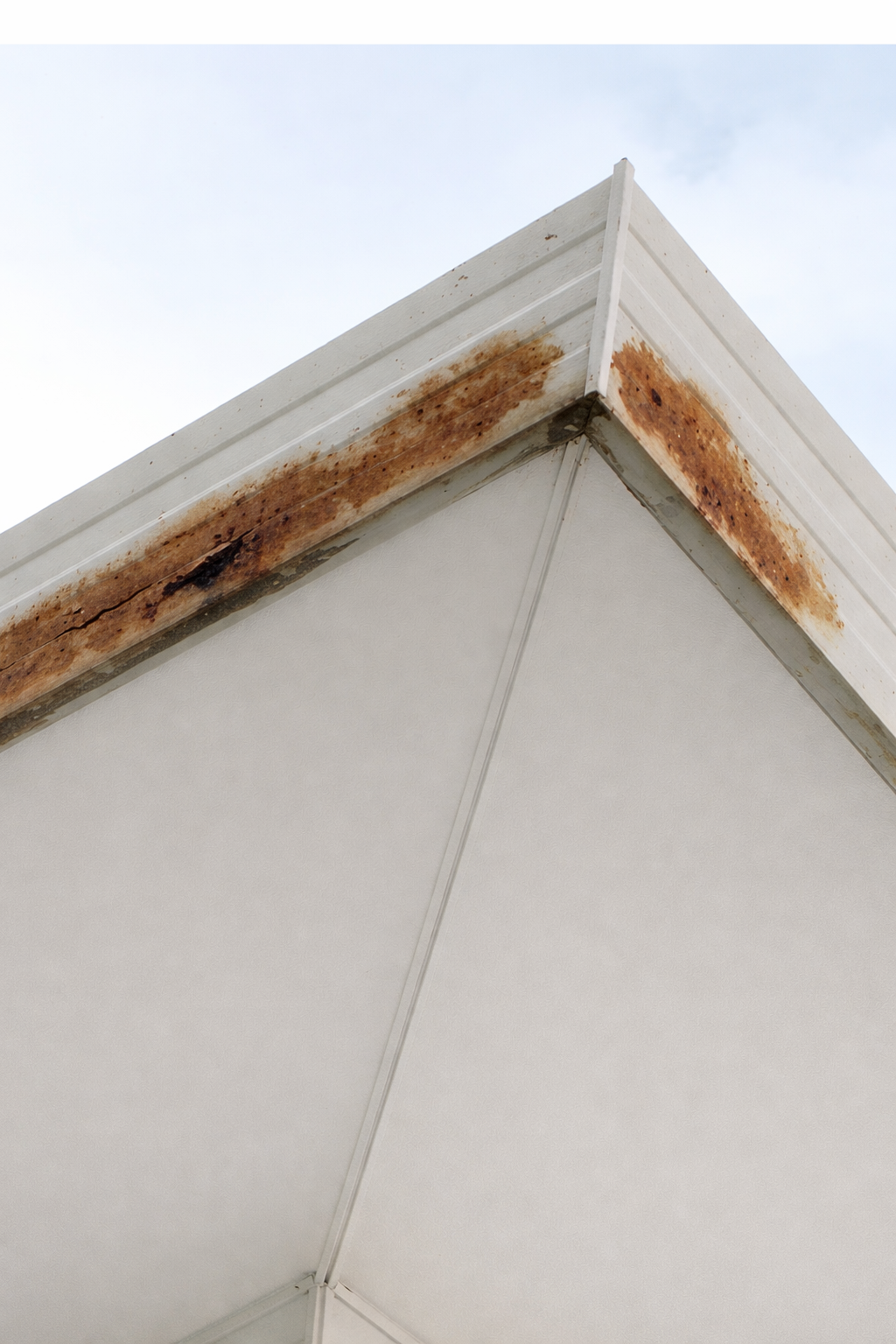 Close-up of the corner of a building with rusted and stained metal trim and white siding, damaged caused by internal gutters.