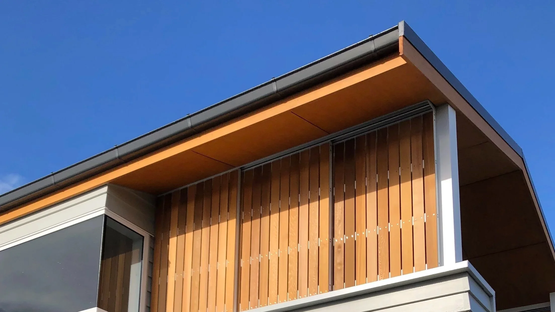 Close-up of a modern building exterior with wooden slats and large glass window under a clear blue sky and modern external gutter system.