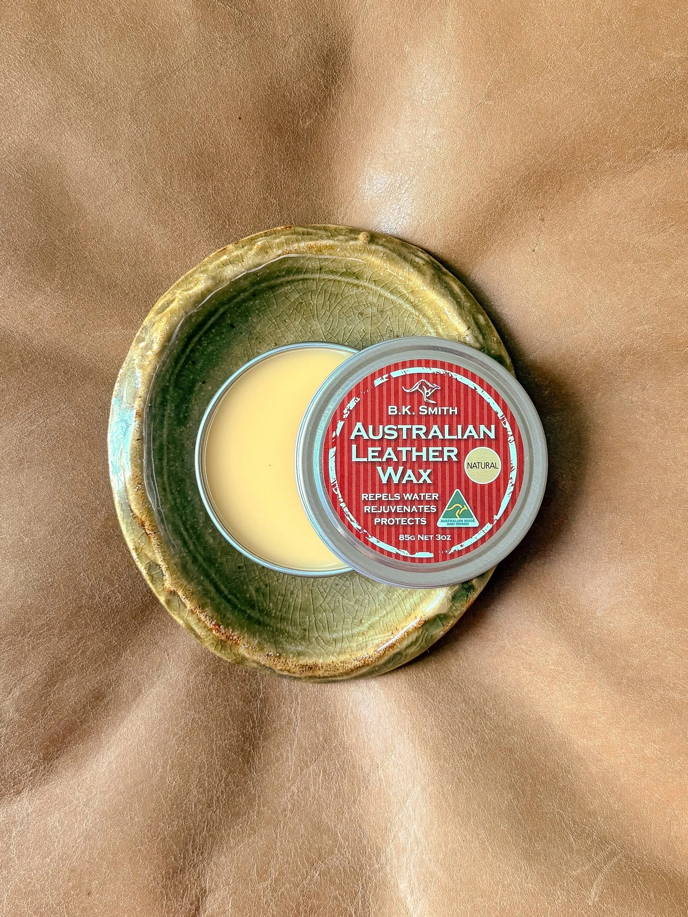 A container of B.K. Smith Australian Leather Wax placed in a decorative green pottery dish with a textured surface, on a light brown leather surface.
