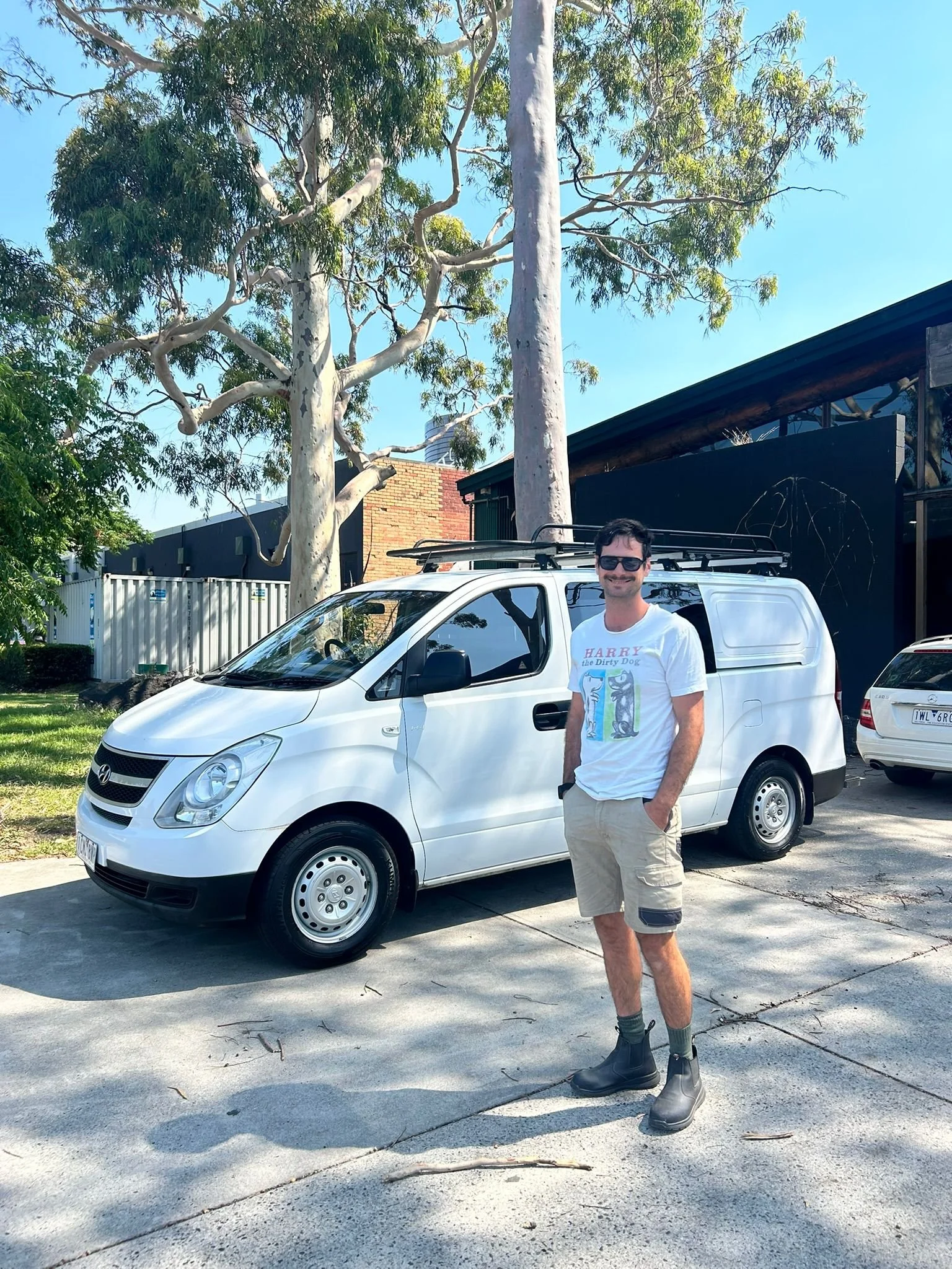 A man standing outdoors in front of a white van, wearing sunglasses, a white T-shirt with graphic print, beige shorts, black socks, and black boots, with large eucalyptus trees and a building in the background under a clear blue sky.