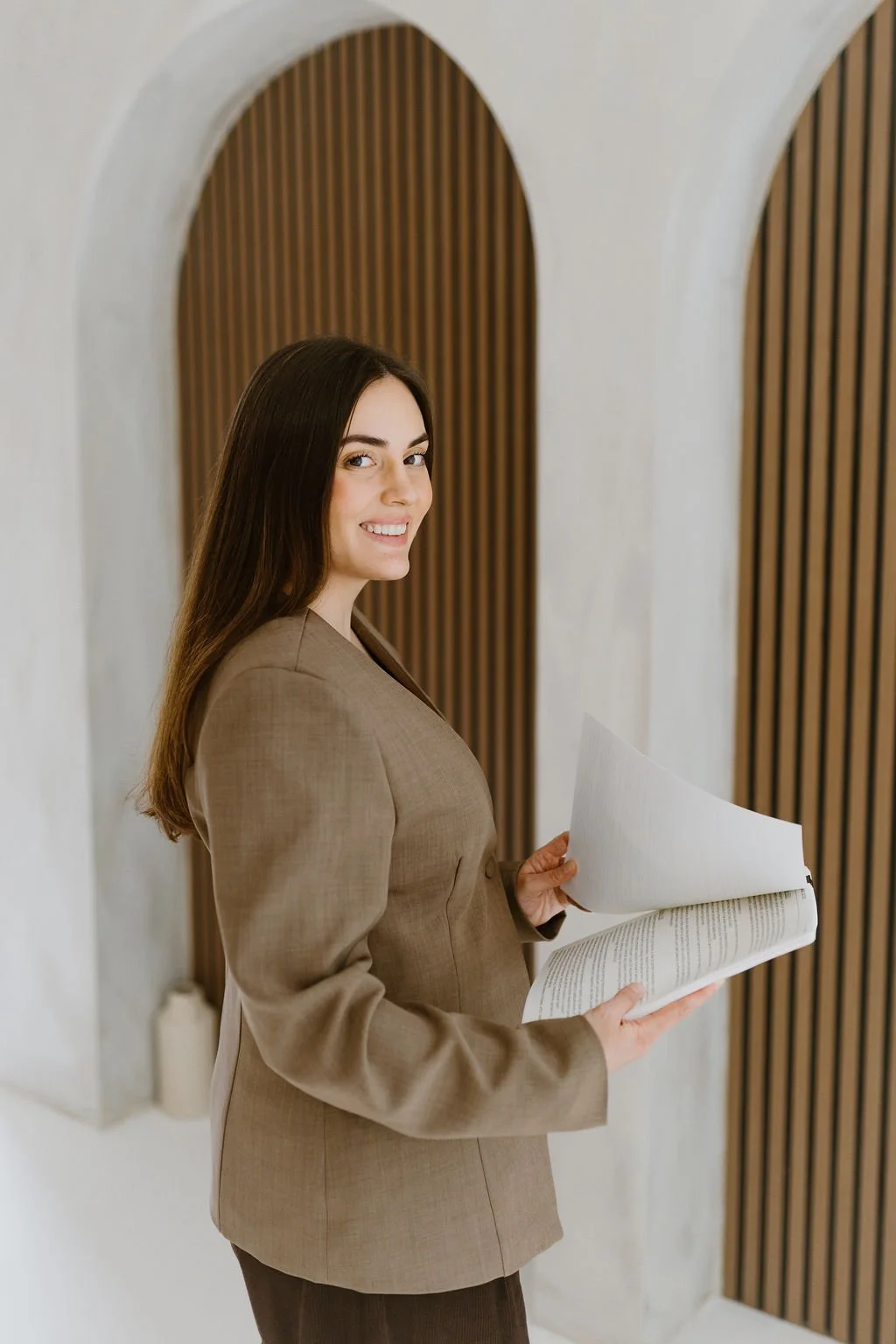 A woman with long dark hair, dressed in a brown suit, is holding and looking at a large book or document, standing in a modern interior with wooden slats and arched architectural features.