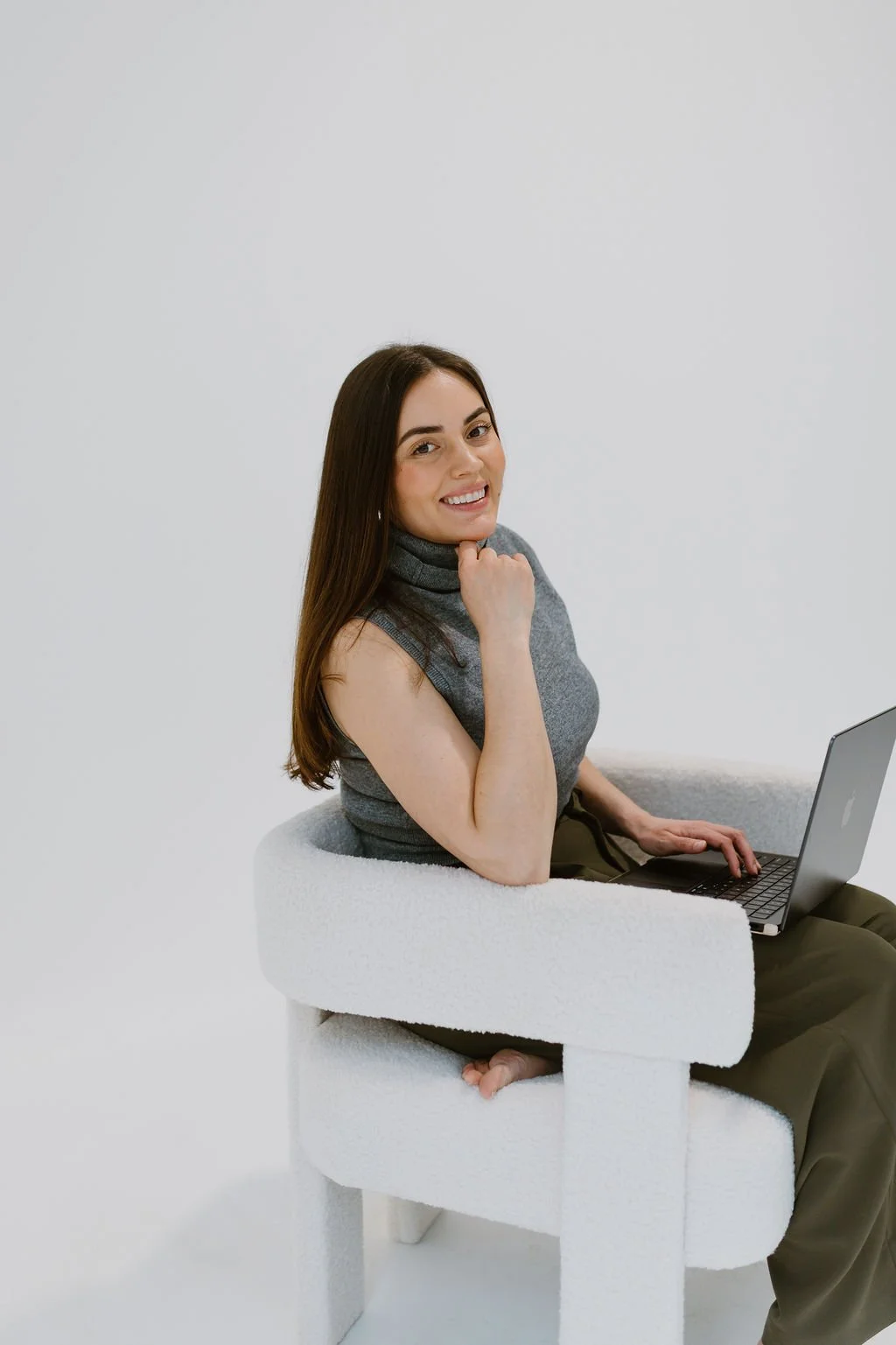 A woman with long brown hair sitting on a white textured chair with a laptop on her lap, smiling and looking at the camera, in front of a plain white background.