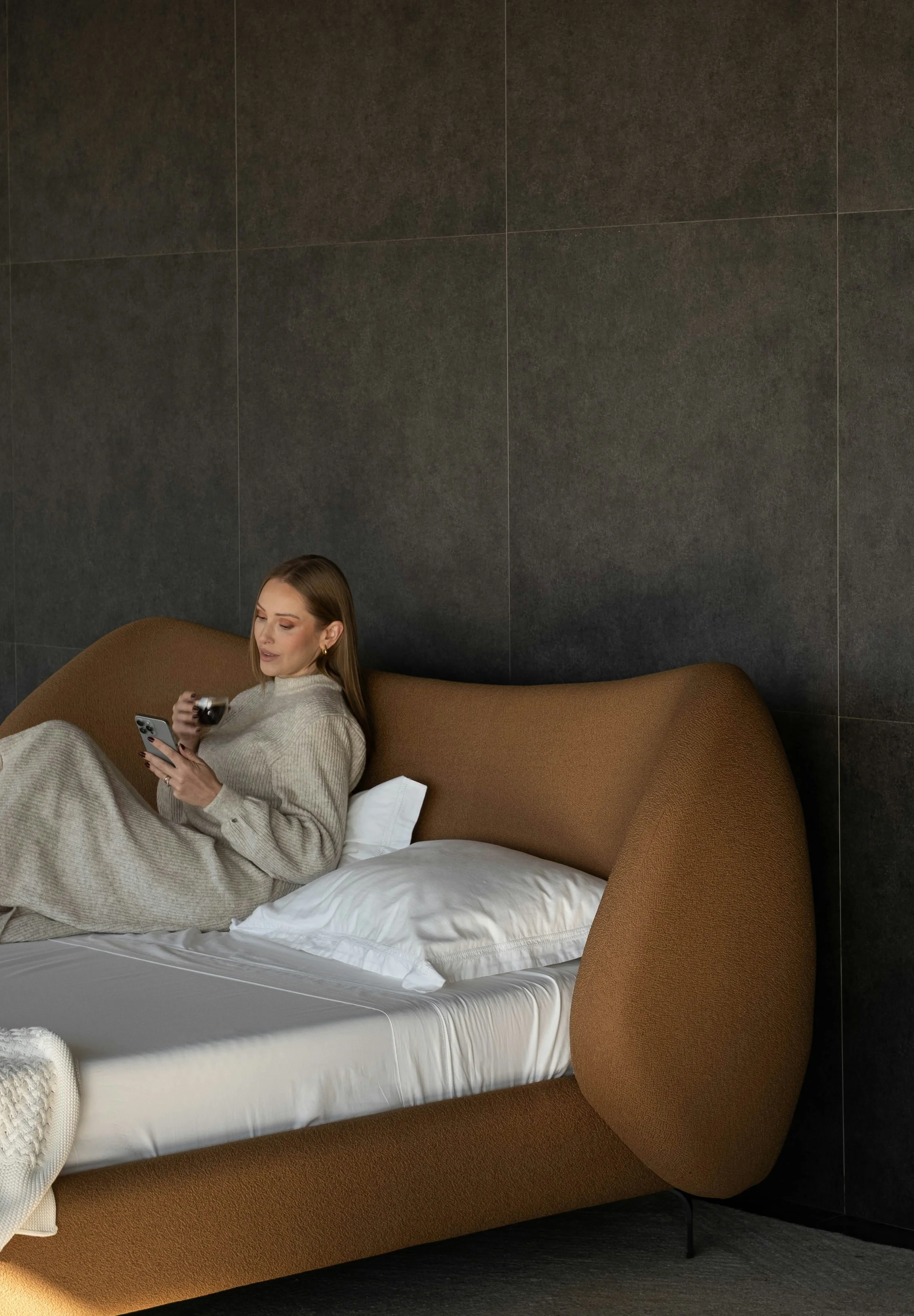 A woman relaxing on a brown modern bed with white sheets and pillows, holding a smartphone and a glass of wine, in a dark modern room.