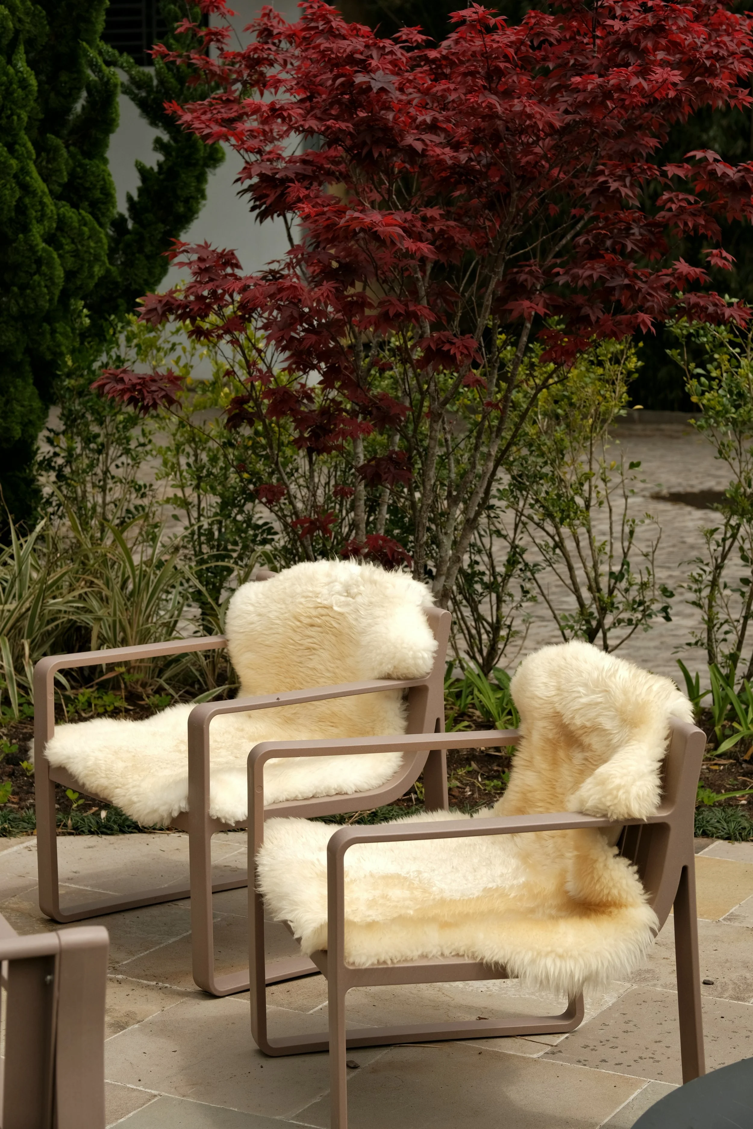 Two modern chairs with beige frames and plush cream-colored faux fur cushions and backrests are placed outdoors on a stone patio, with red and green foliage in the background.
