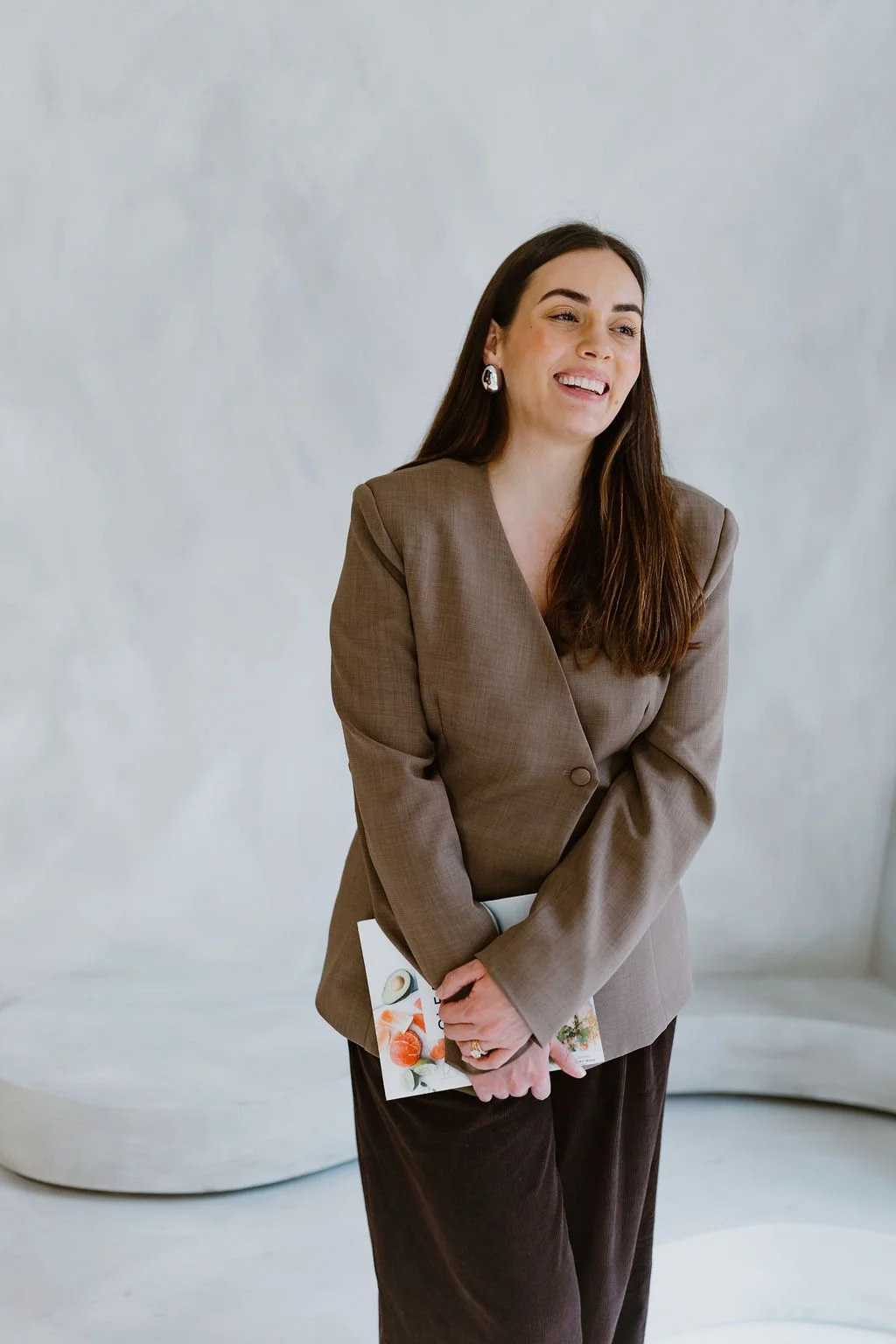 A woman with long dark hair wearing a tan blazer and black pants, holding a magazine or booklet, smiling in front of a plain white background.