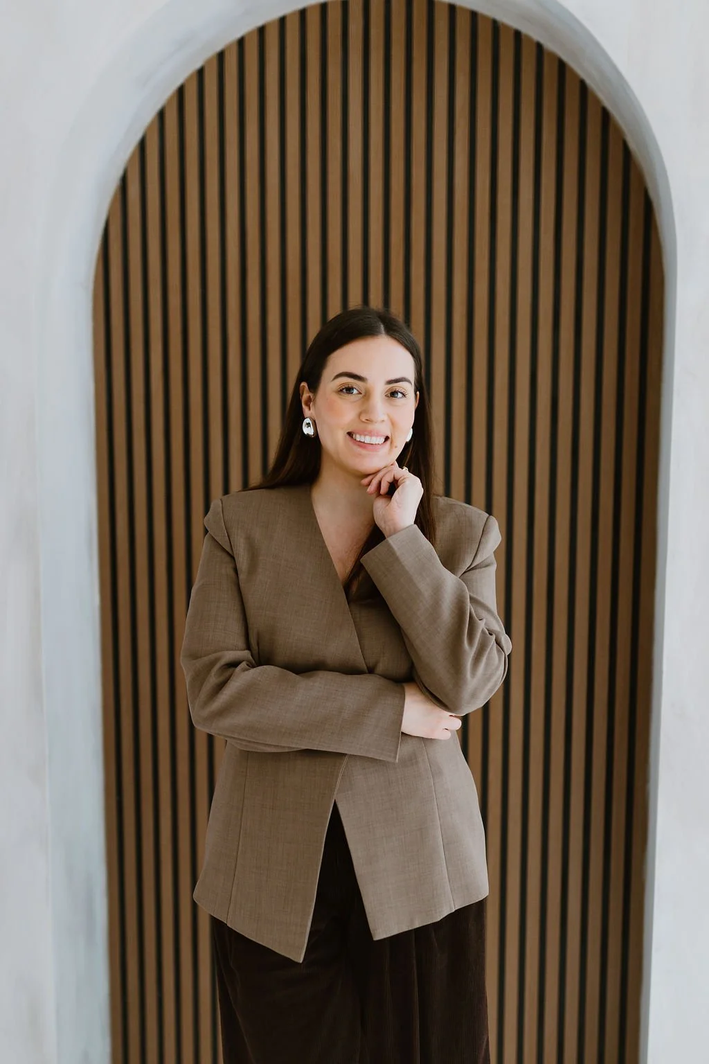 A woman with long dark hair, wearing a tan blazer and dark pants, standing in front of a wooden slat wall inside an arched doorway, smiling and looking at the camera.