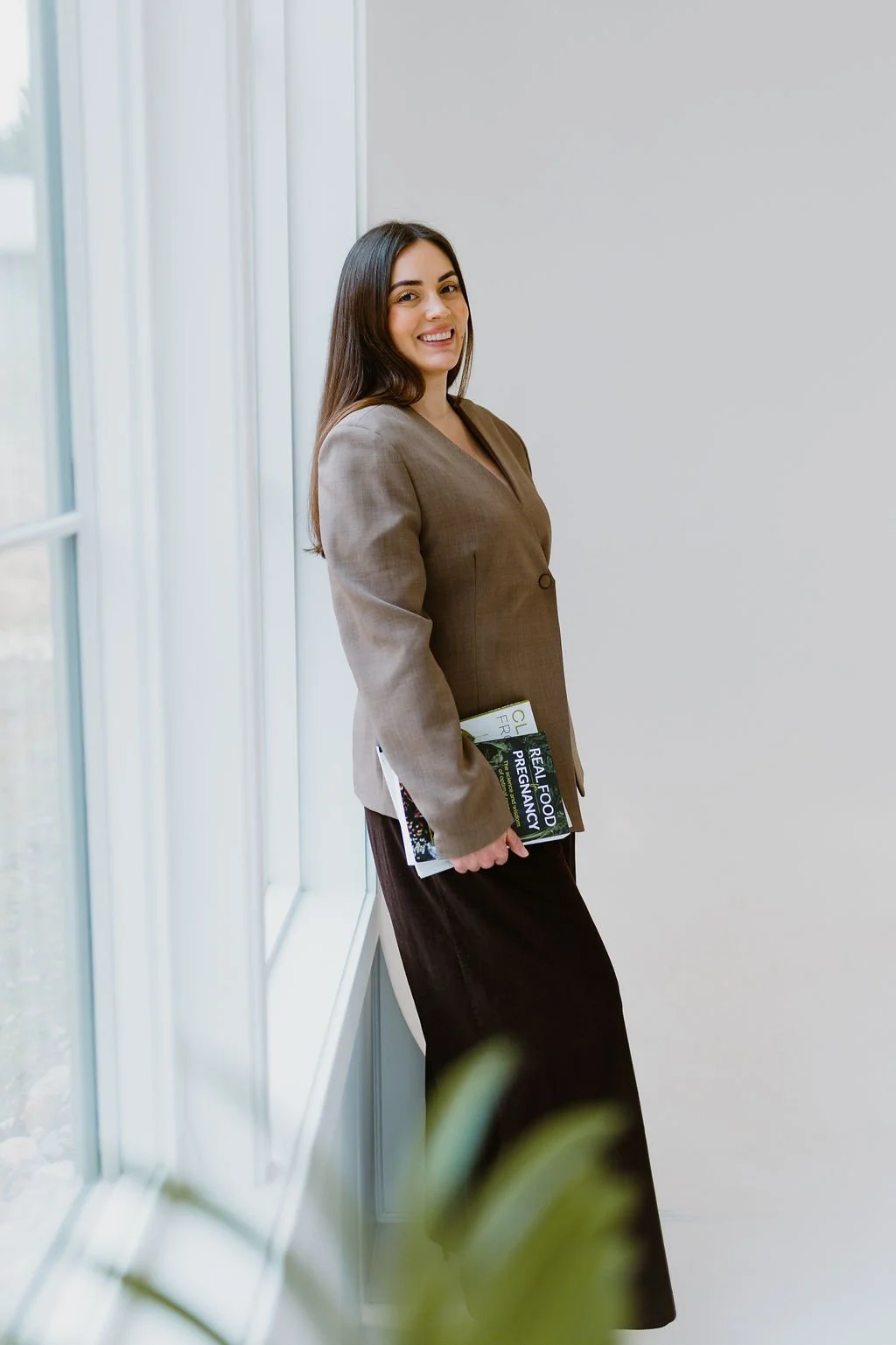 A woman with long dark hair smiling while holding magazines, standing by a large window in a bright room.