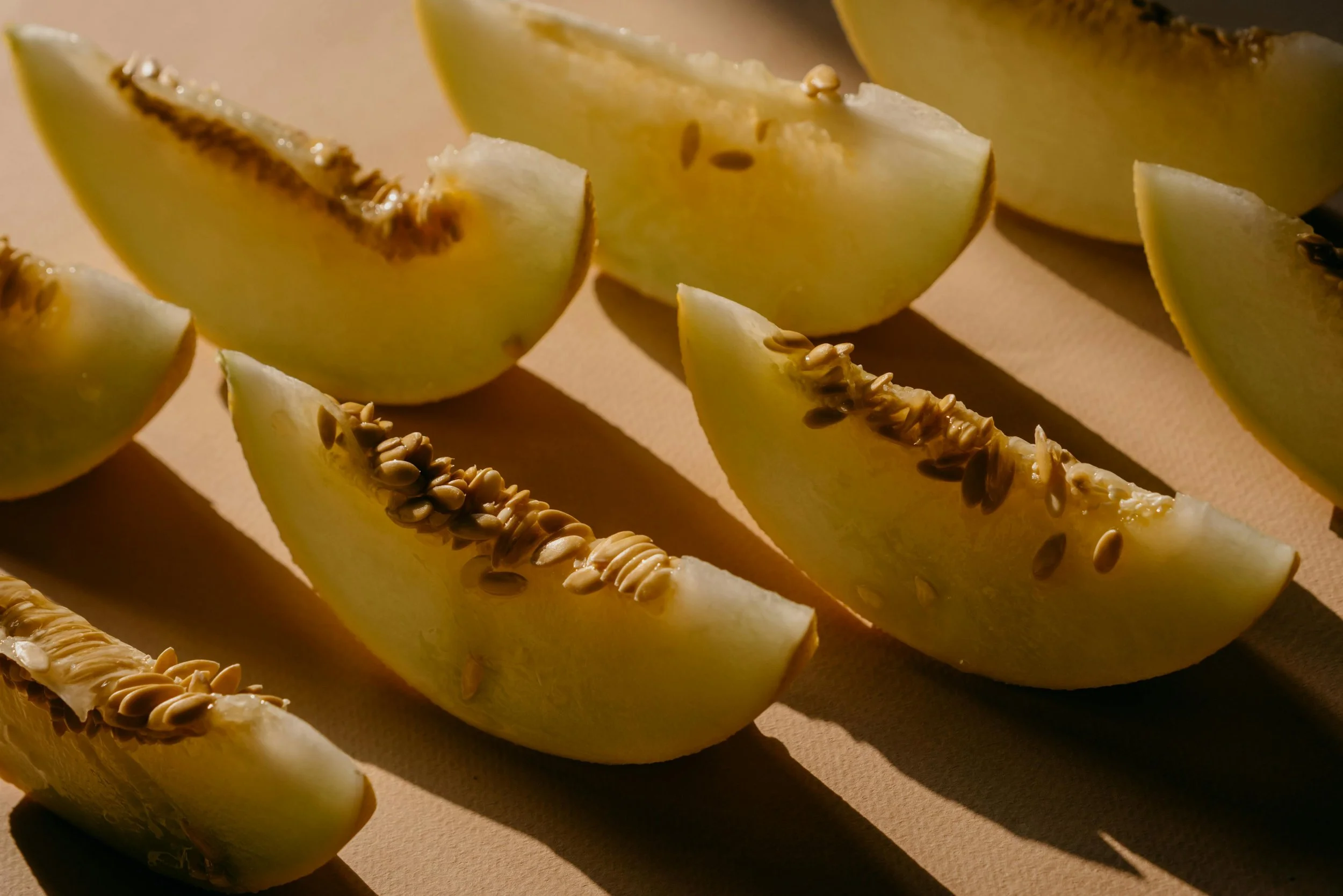 Slices of yellow melon with seeds on a light surface, casting shadows.