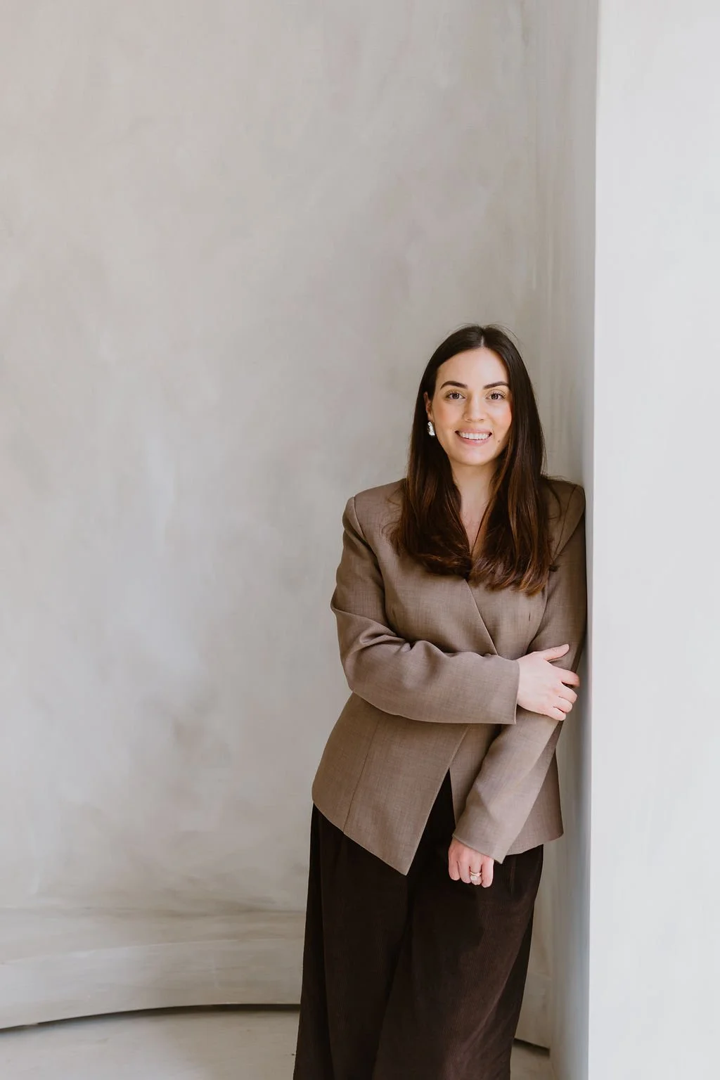 A woman with long brown hair, wearing a beige blazer, smiling and standing with one arm crossed in front of her, leaning against a white wall.