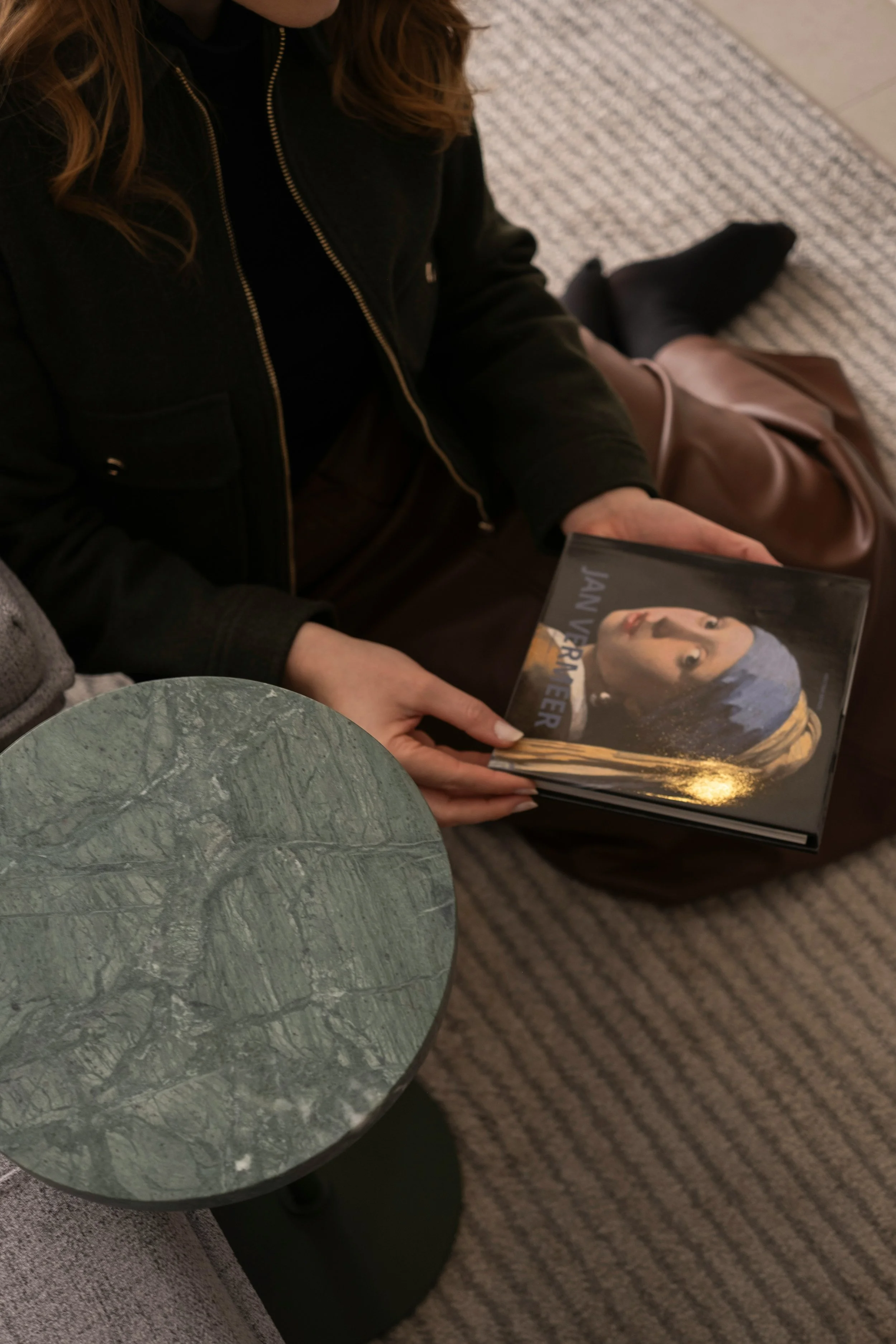 Person holding a hardcover book titled 'Jane Vereeer' with a portrait of a woman with blue hair on the cover, sitting on a striped carpet next to a small round marble-topped table.