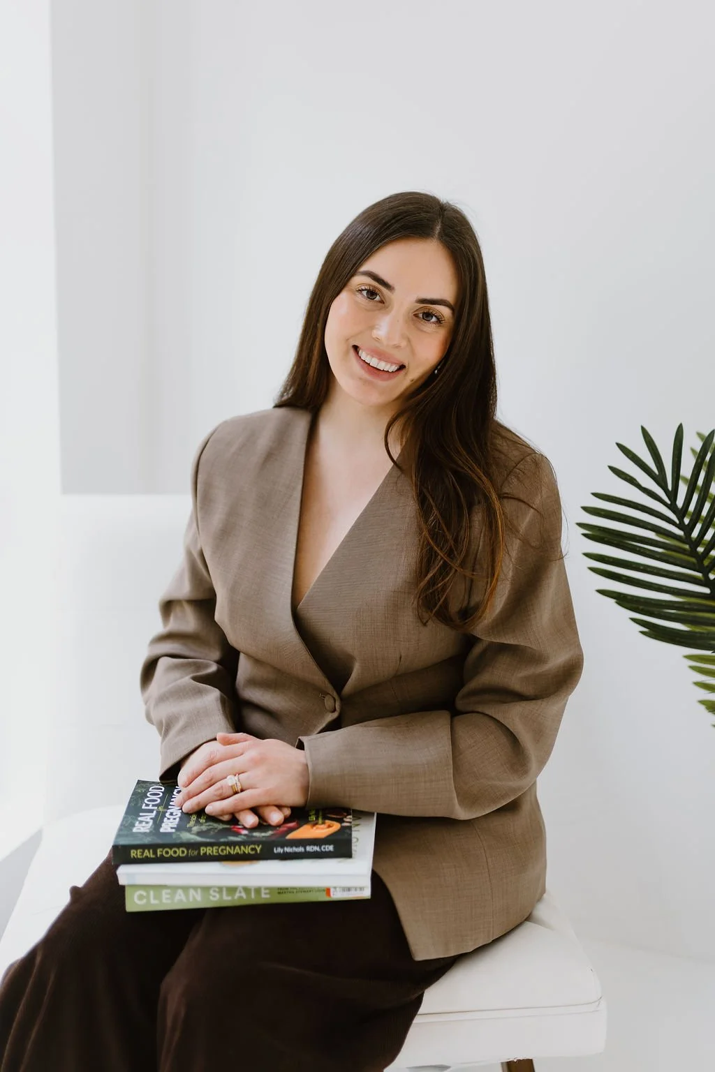 A young woman with long brown hair, wearing a brown blazer, sitting on a white bench, holding two books, smiling at the camera, with a green plant on her right side.