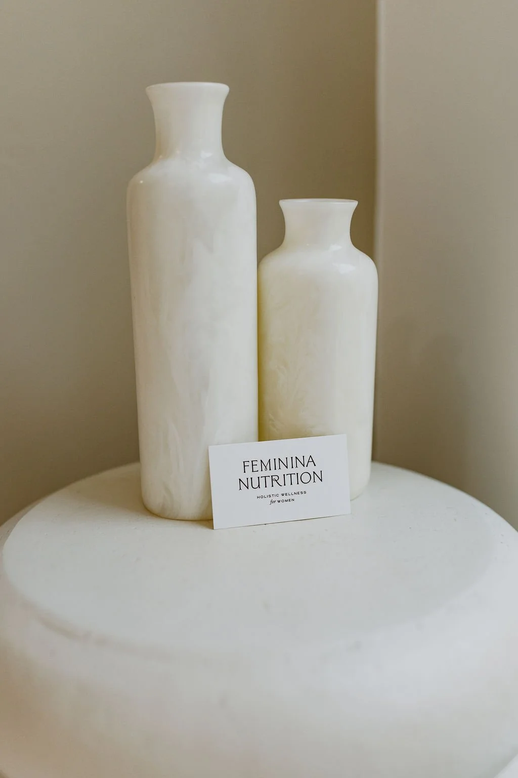 Two white marble vases of different heights on a round white table, with a small white card reading "Feminina Nutrition, Holistic Wellness for Women" placed in front of them.
