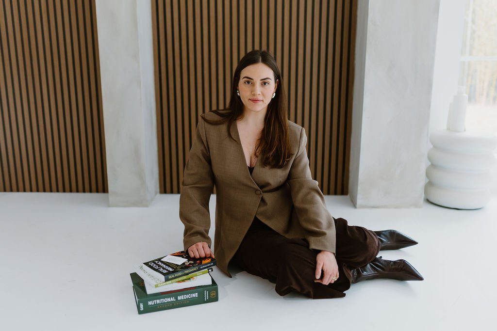 A woman with long brown hair sits on the floor against a wooden slatted wall, with books stacked in front of her, wearing a brown blazer and dark pants.