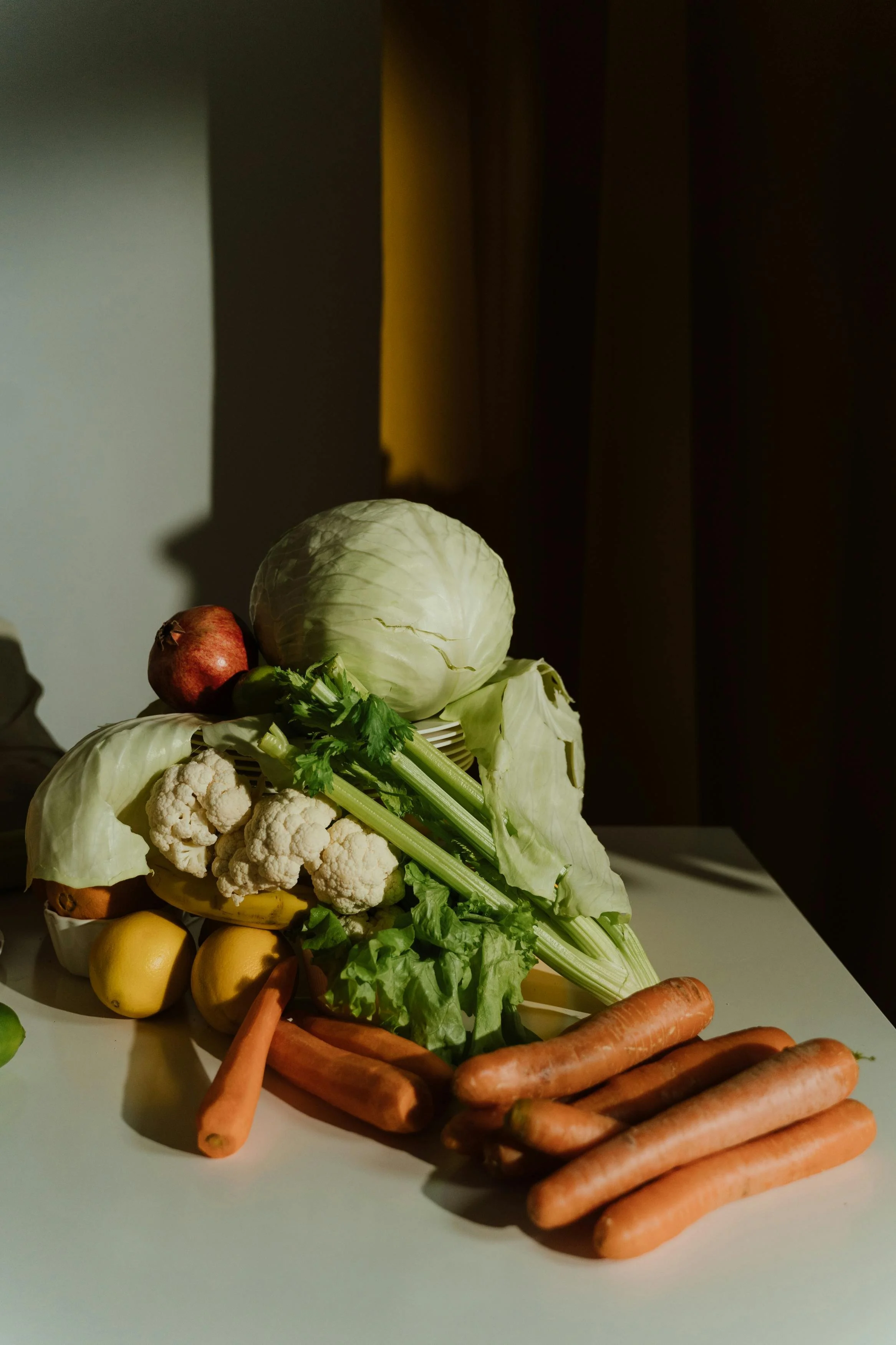 Assorted vegetables including carrots, cauliflower, cabbage, celery, apples, and pomegranate on a white surface in front of a dark curtain with light coming from the left.