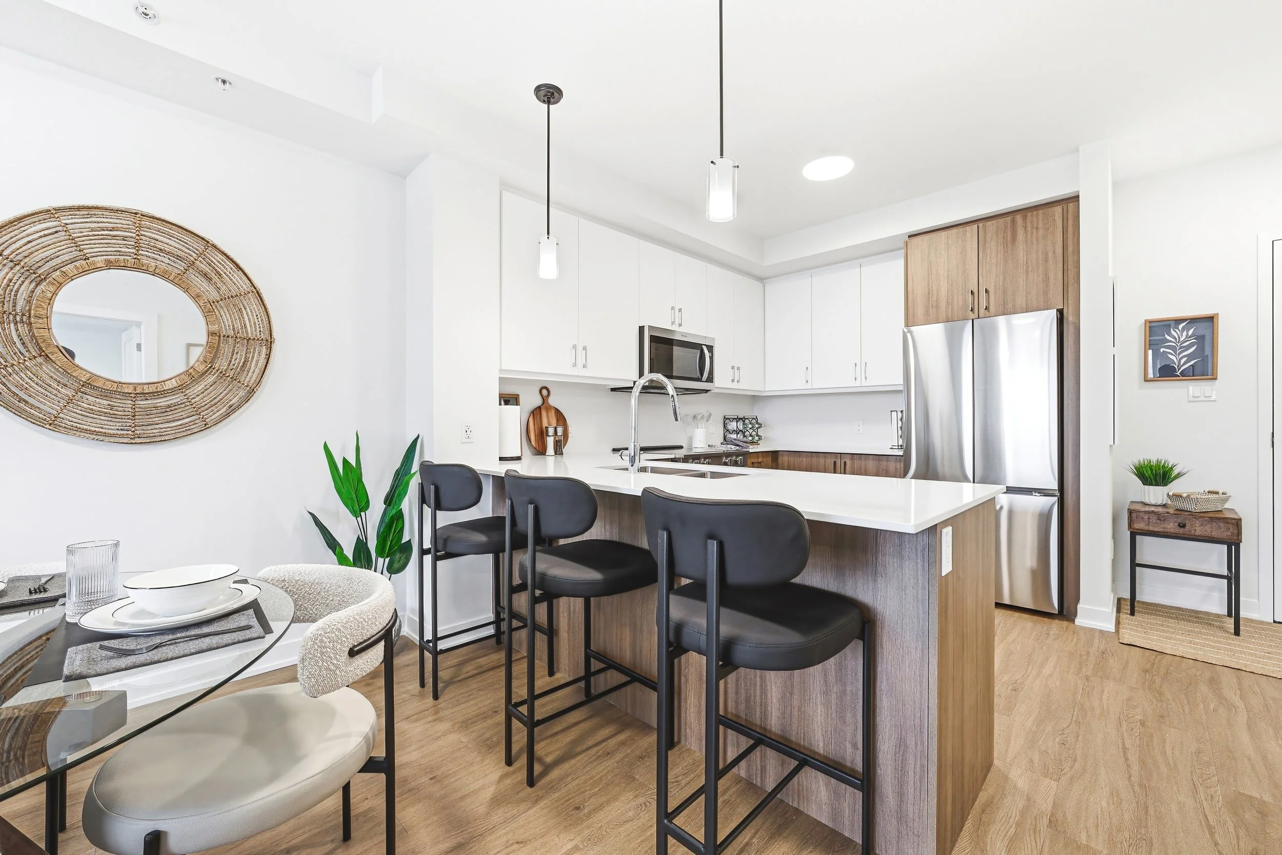 Modern kitchen with white cabinets, stainless steel refrigerator, black bar stools, and a dining area with a glass table and beige chairs.