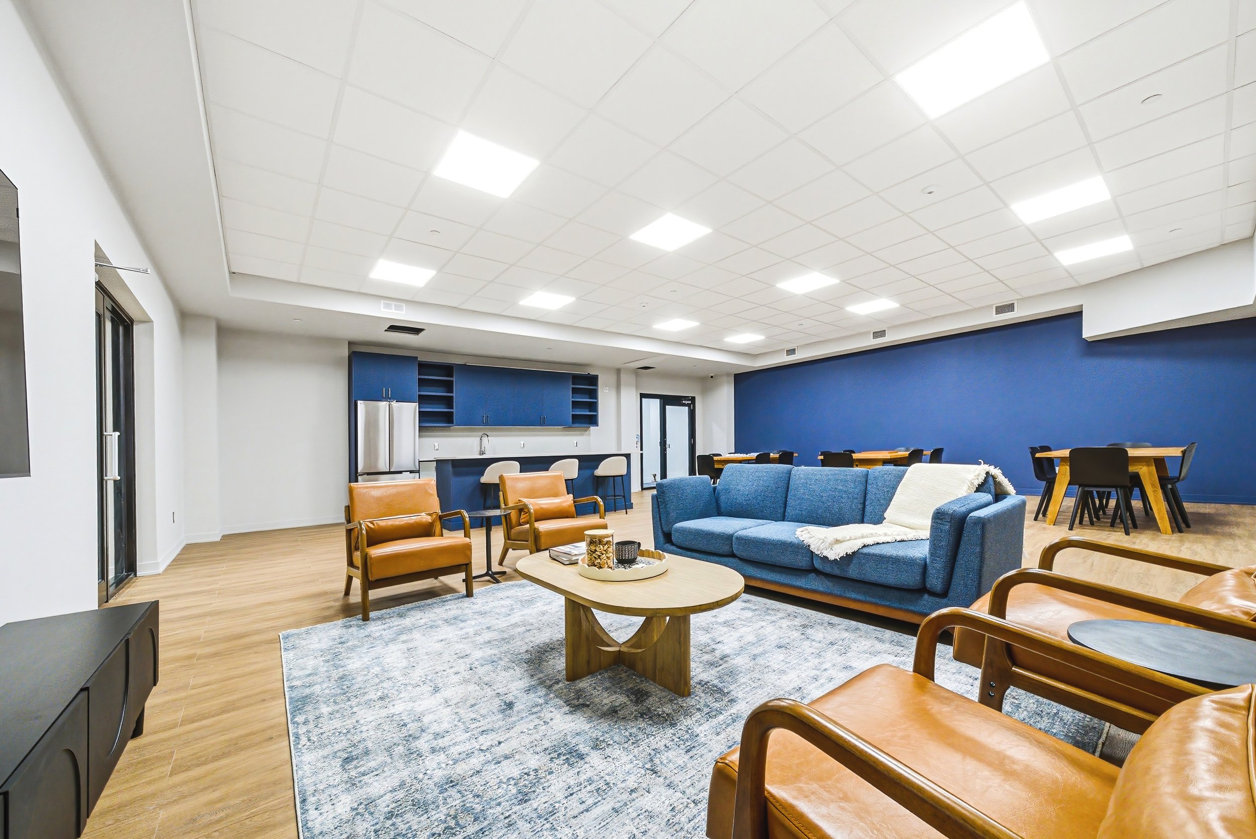 A spacious lounge area with blue and beige couches, wooden chairs, a central oval wooden coffee table, a rug, and a kitchenette with blue cabinets and a stainless steel refrigerator. The ceiling has white tiles with rectangular fluorescent lights, and the walls are painted white and blue.