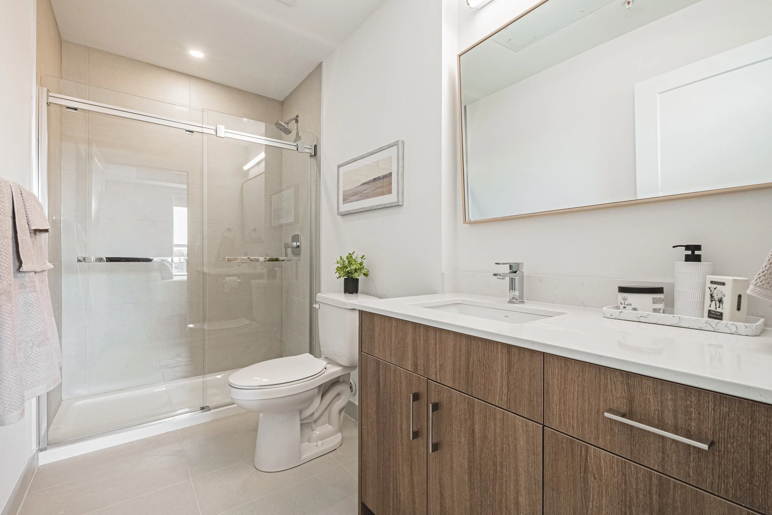 A modern bathroom with a glass shower enclosure, white toilet, wooden vanity with white countertop, large mirror, some artwork on the wall, and decorative items like a potted plant and soap dispenser.