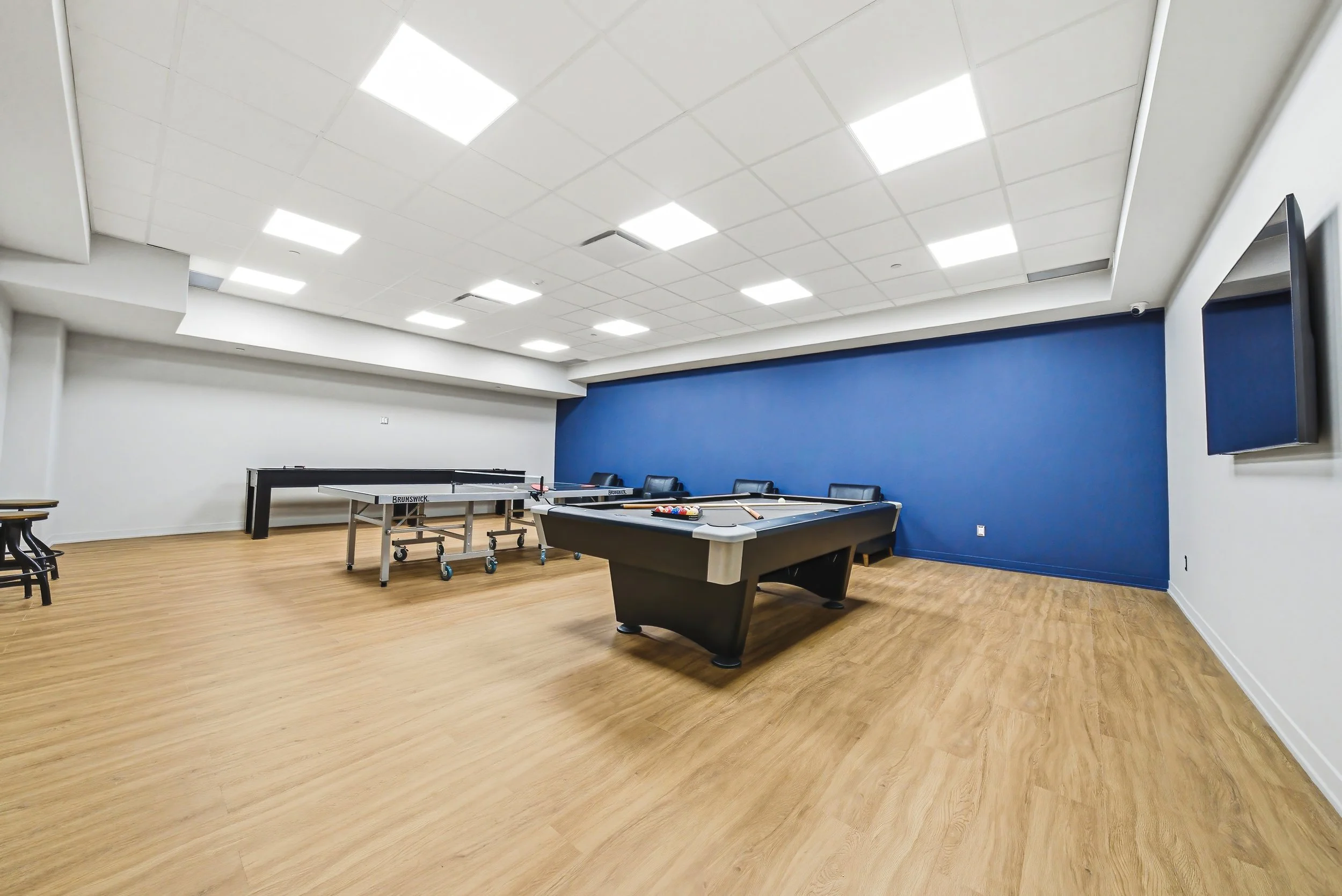 An empty indoor recreation room with a ping pong table and a pool table, white ceiling with square lights, white and blue walls, and light wood floor.
