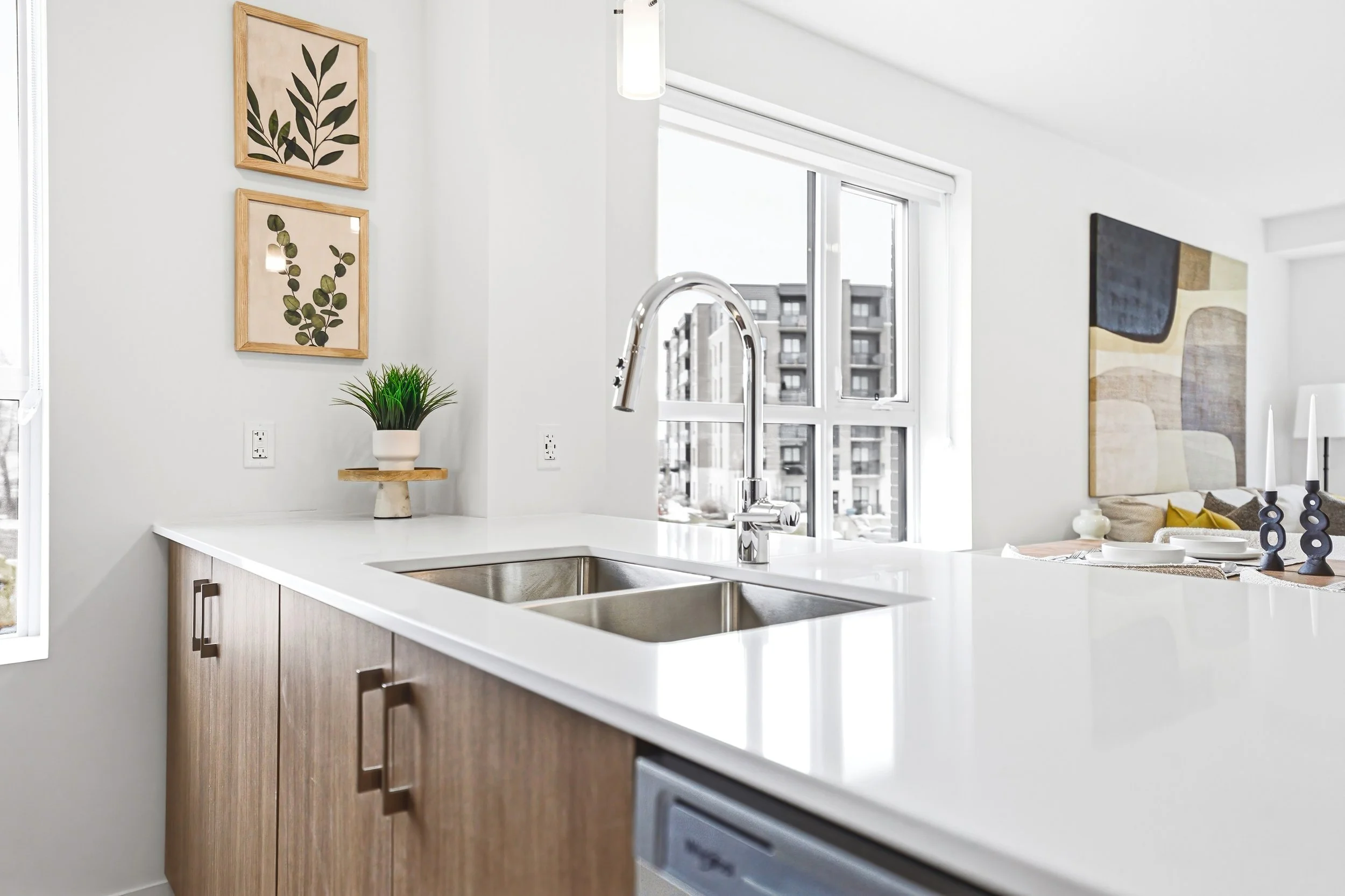 Modern kitchen with a white countertop, stainless steel sink, wooden cabinets, a potted plant, and artwork on the wall, with windows showing an urban building outside.