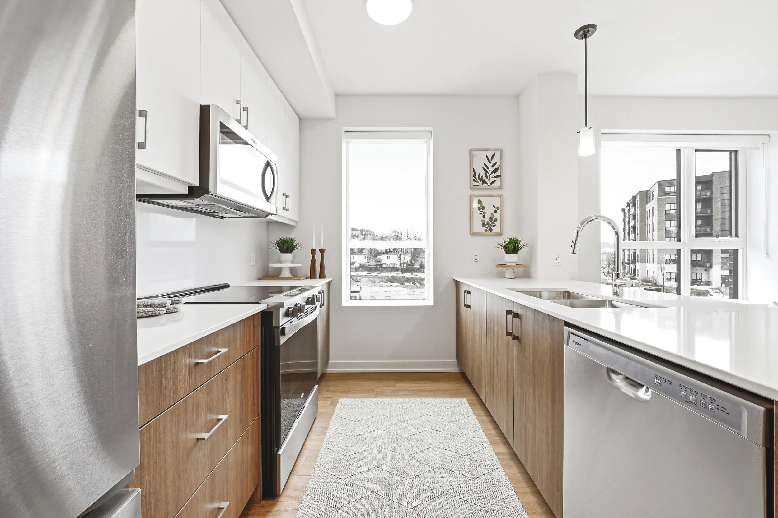 Modern kitchen with white upper cabinets, wooden lower cabinets, stainless steel appliances, a dishwater, and a white countertop. There are windows letting in natural light, decorative plants, and framed wall art.