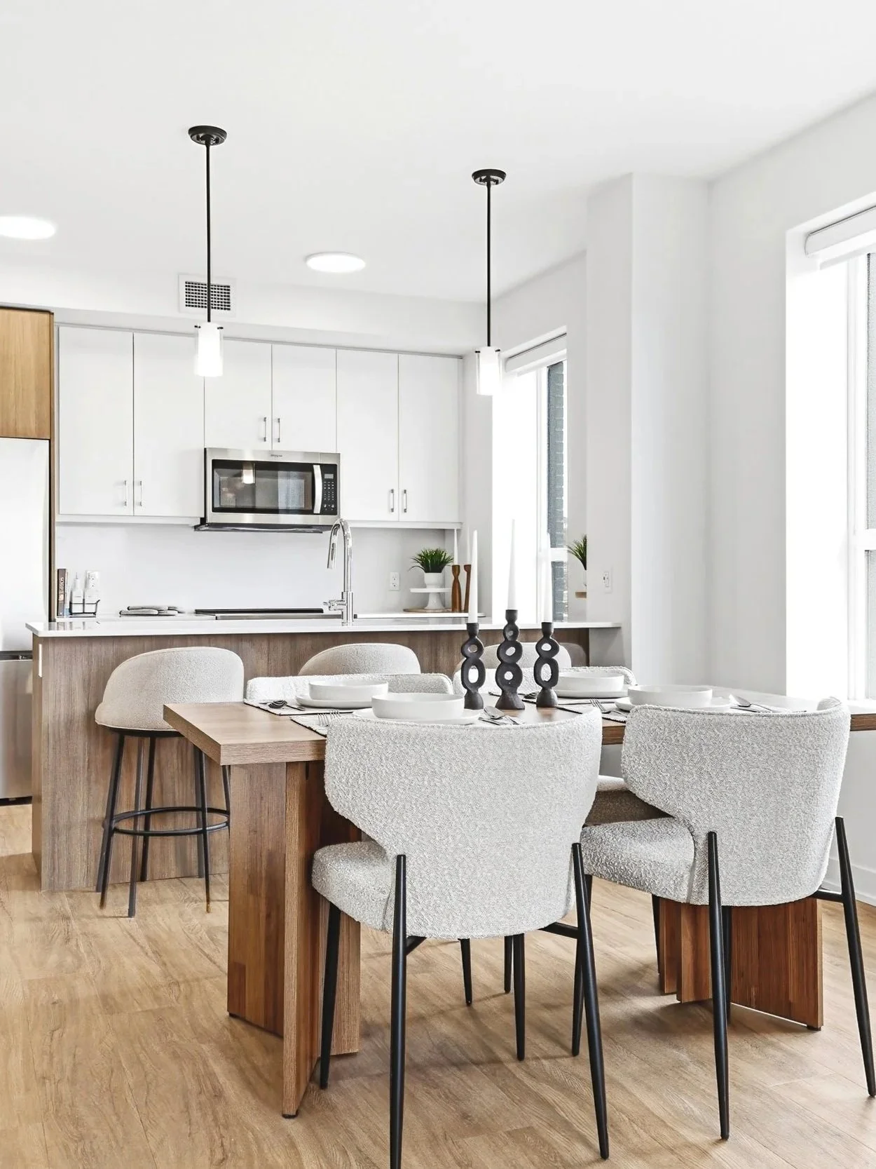 Modern kitchen and dining area with light wood flooring, white cabinets, stainless steel microwave, and a wooden dining table surrounded by six beige upholstered chairs with black legs. The table has white bowls and black decorative candle holders.